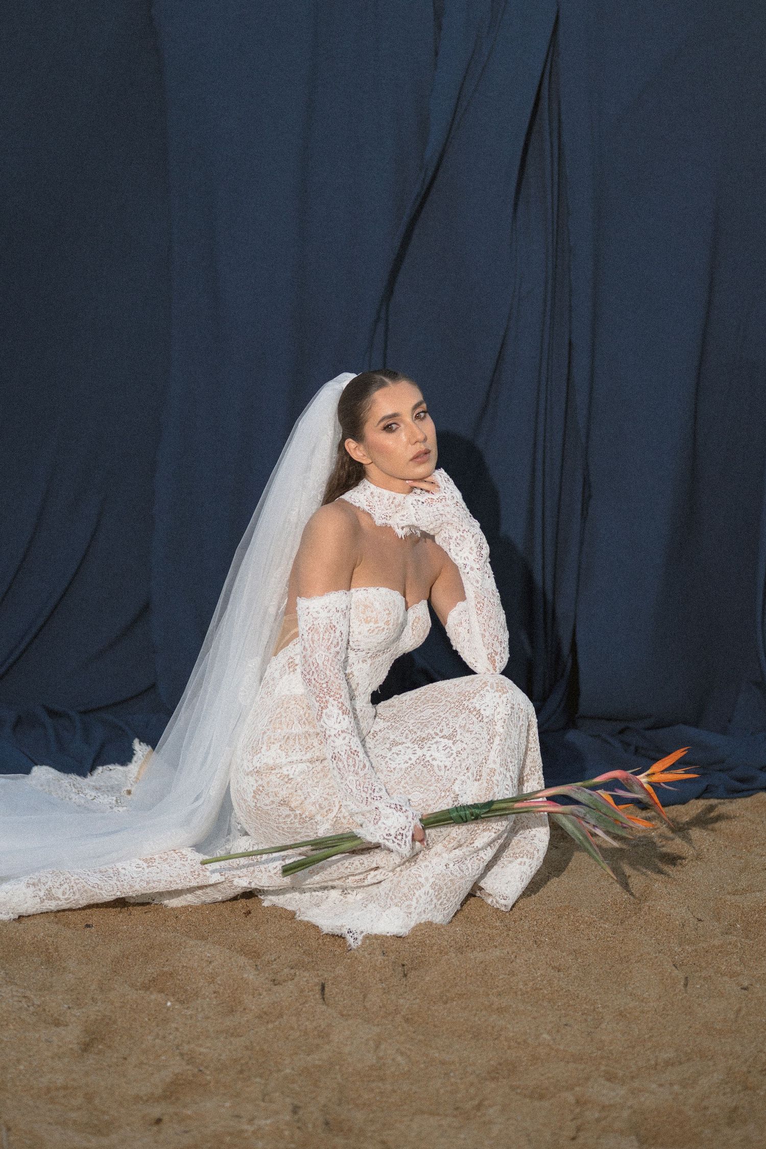 Bride in a lace gown with long veil sits on sand holding a bird of paradise flower against a deep blue backdrop.