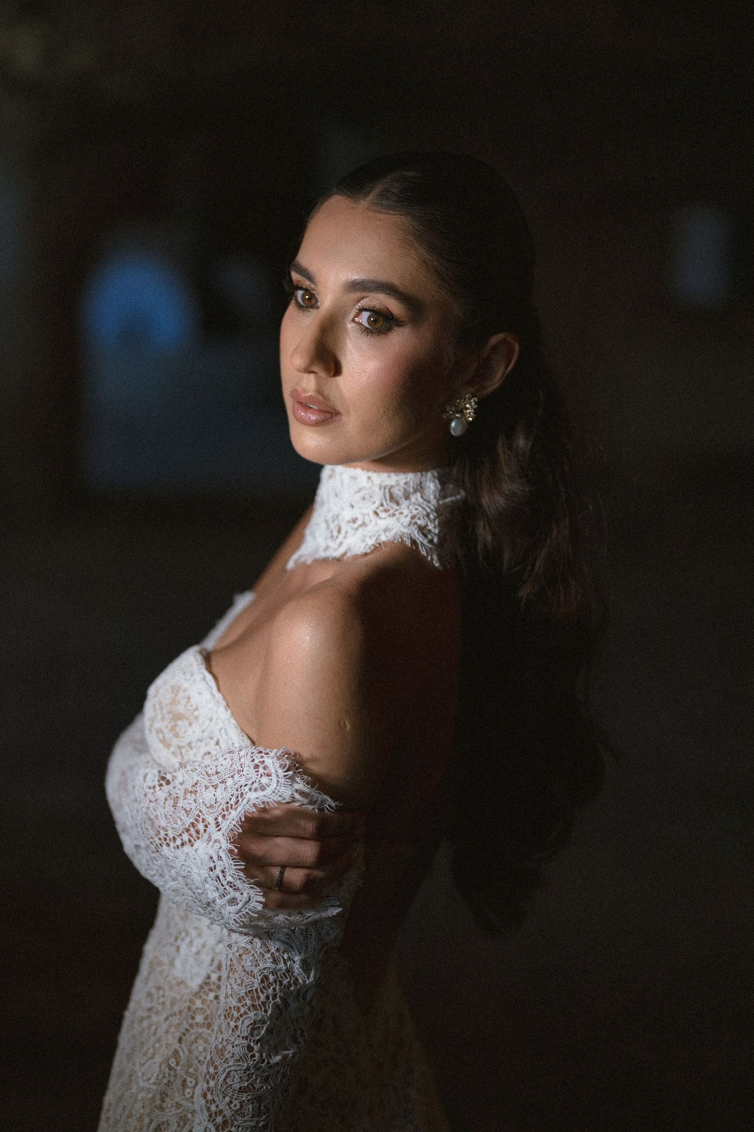 Bride in a lace wedding gown with a high collar looks over her shoulder in soft, moody dramatic lighting.