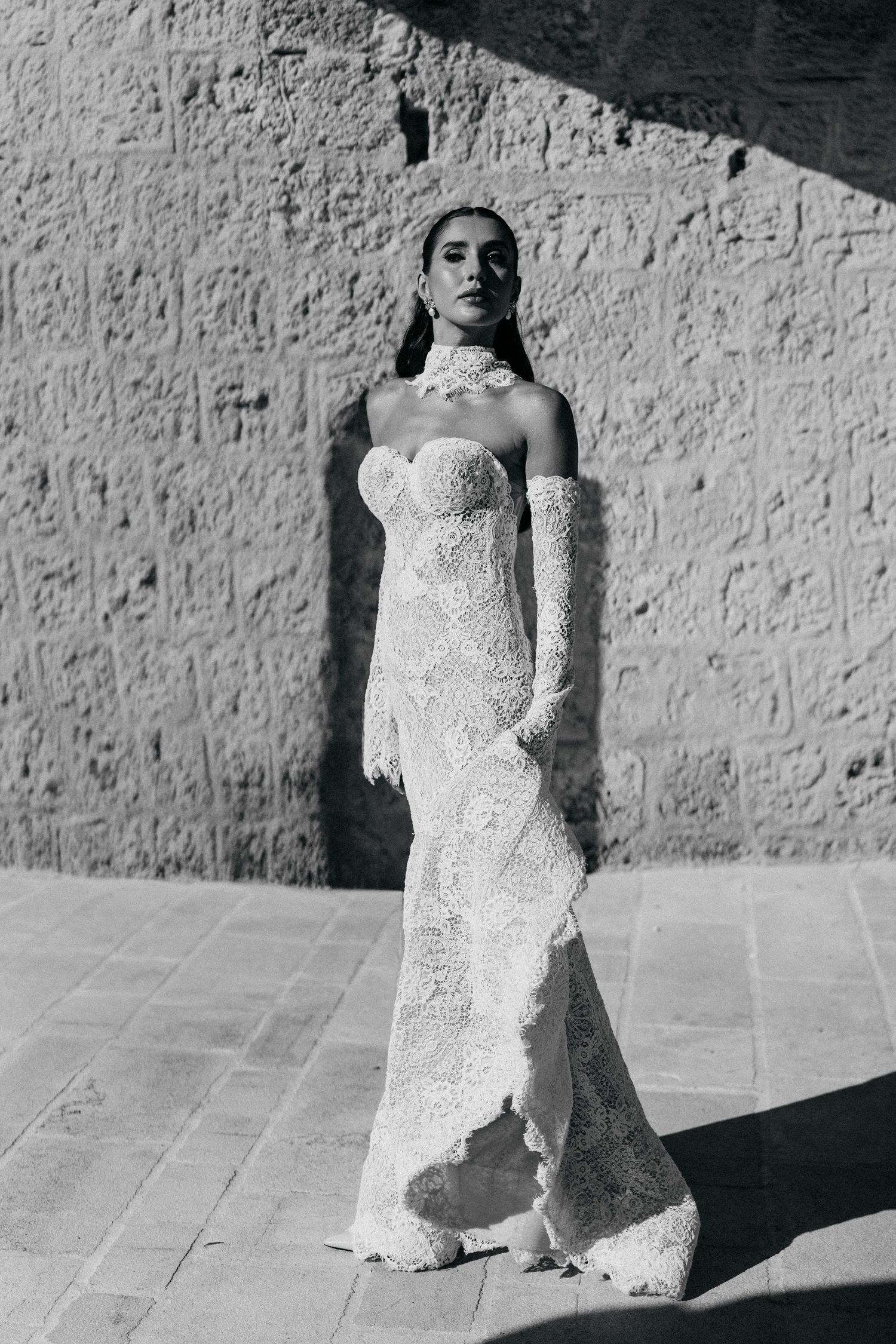 Black and white photo of a bride in a strapless lace wedding gown with a high collar and long gloves, standing in dramatic sunlight.