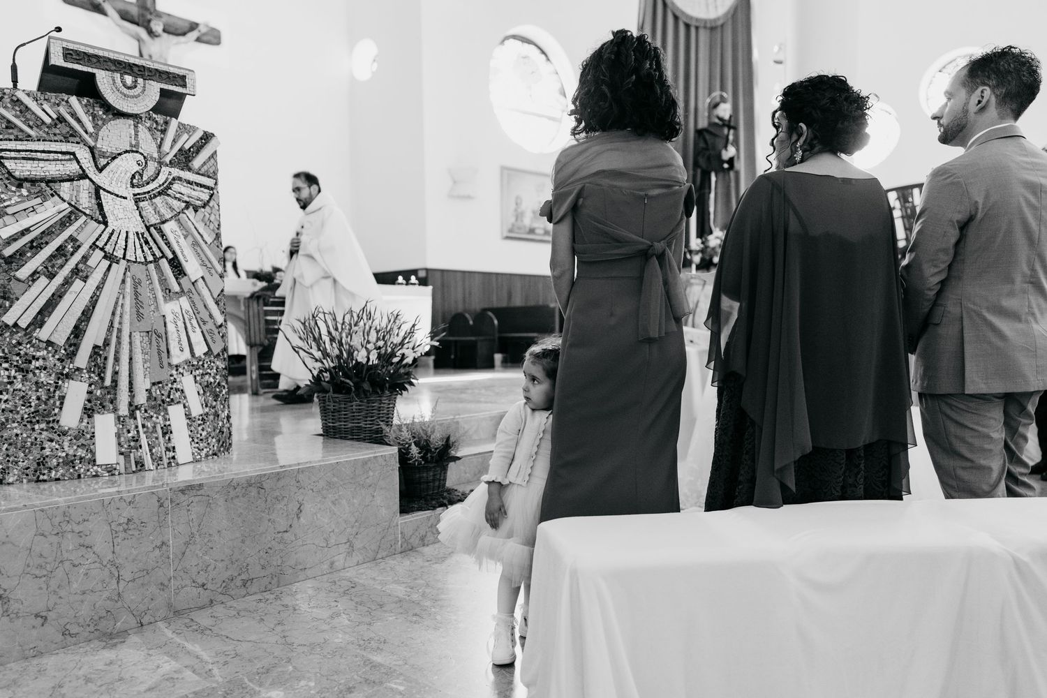 Black and white photo of wedding guests standing during a church ceremony with decorative mosaic altar.