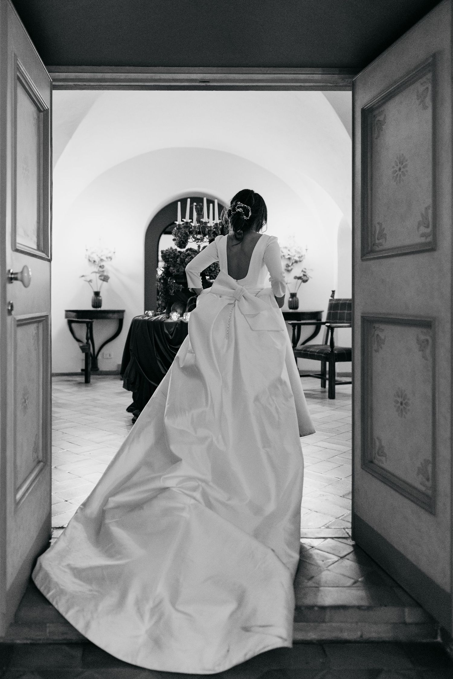 A bride in a long white gown with a bow stands in a doorway in black and white.