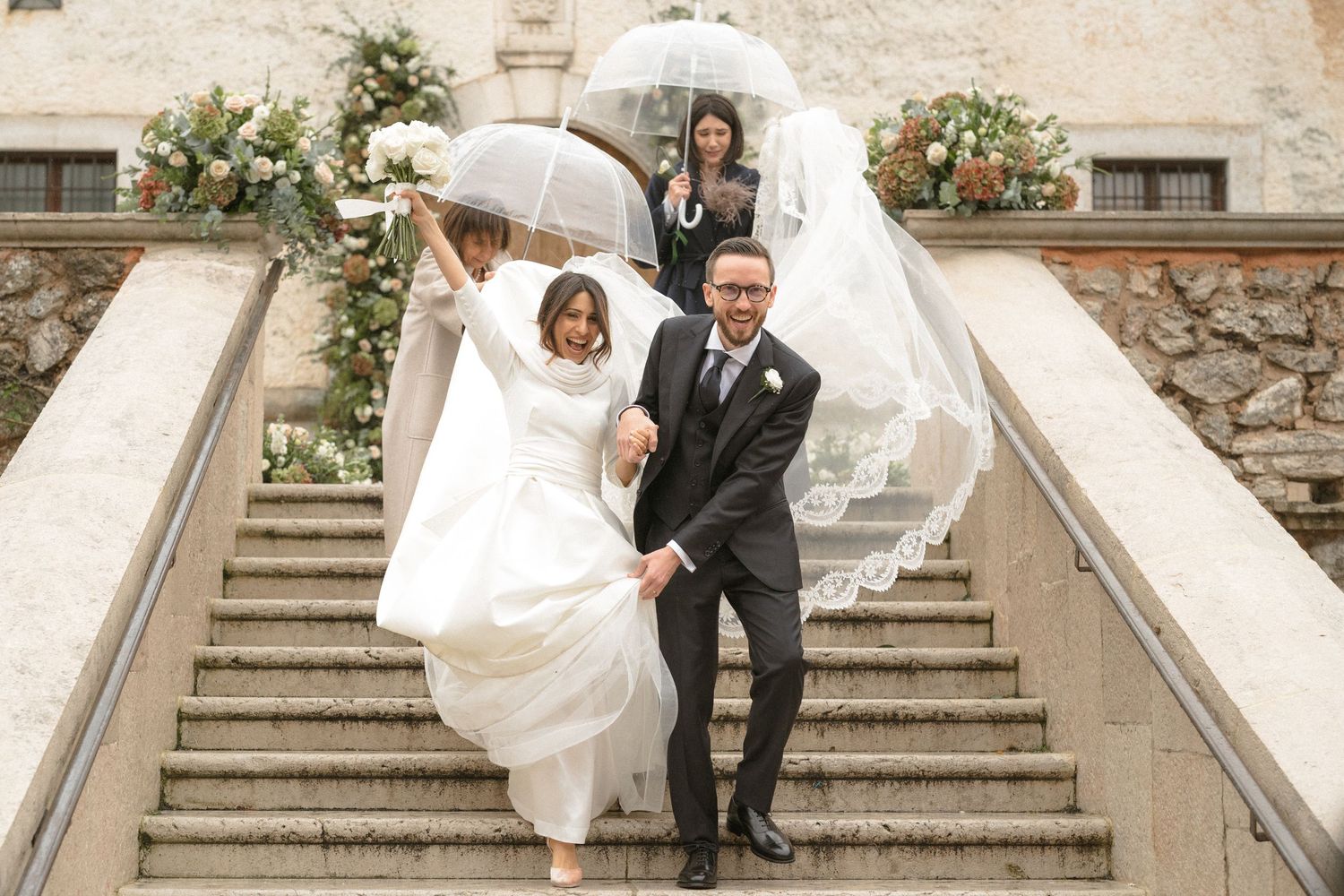 Joyful newlyweds descending stone steps with transparent umbrella as bride lifts wedding gown.