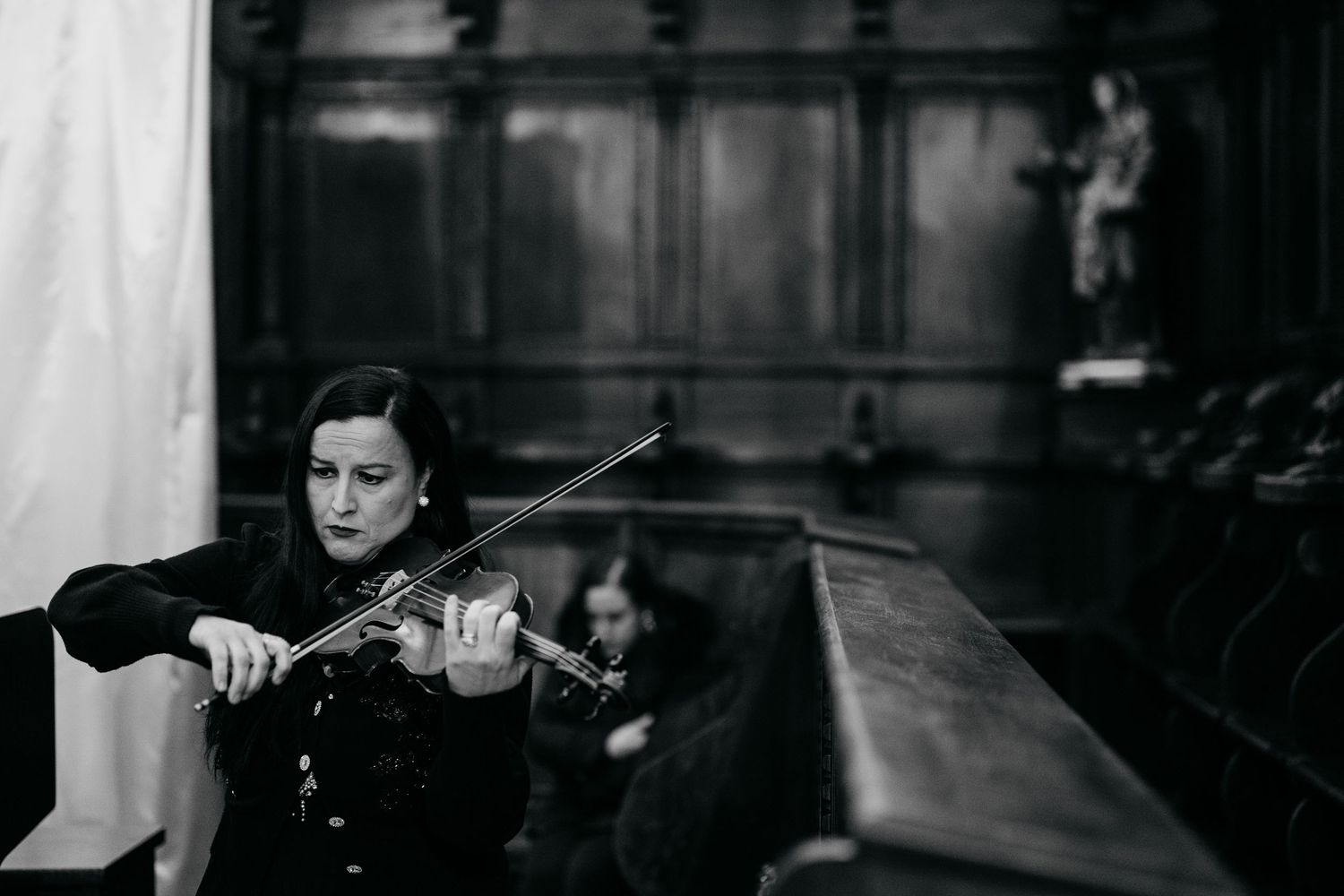 A woman in a black dress plays violin in a grand wood-paneled hall during a wedding ceremony.