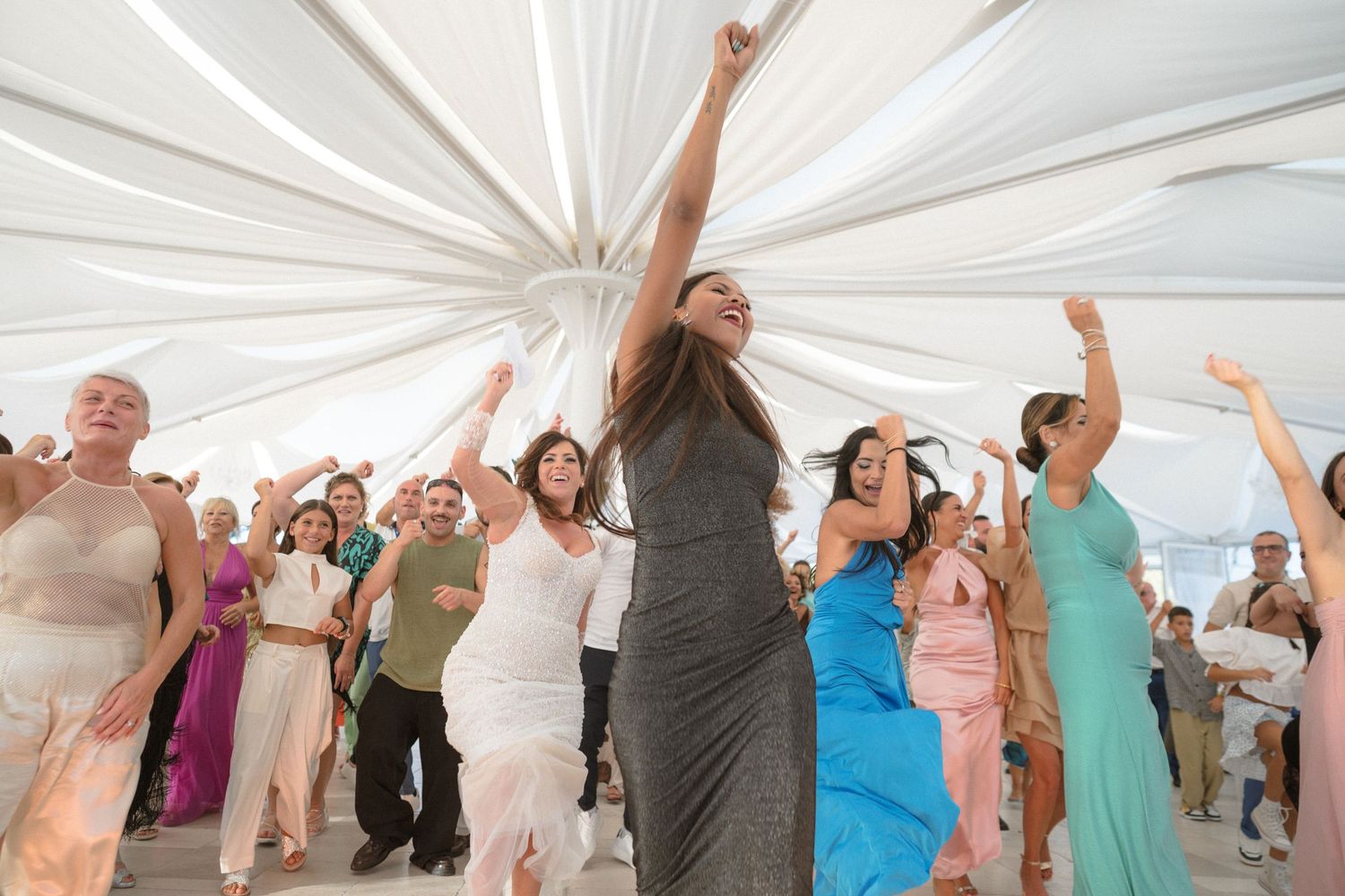 Wedding guests dancing and celebrating under a white tent canopy at a reception.