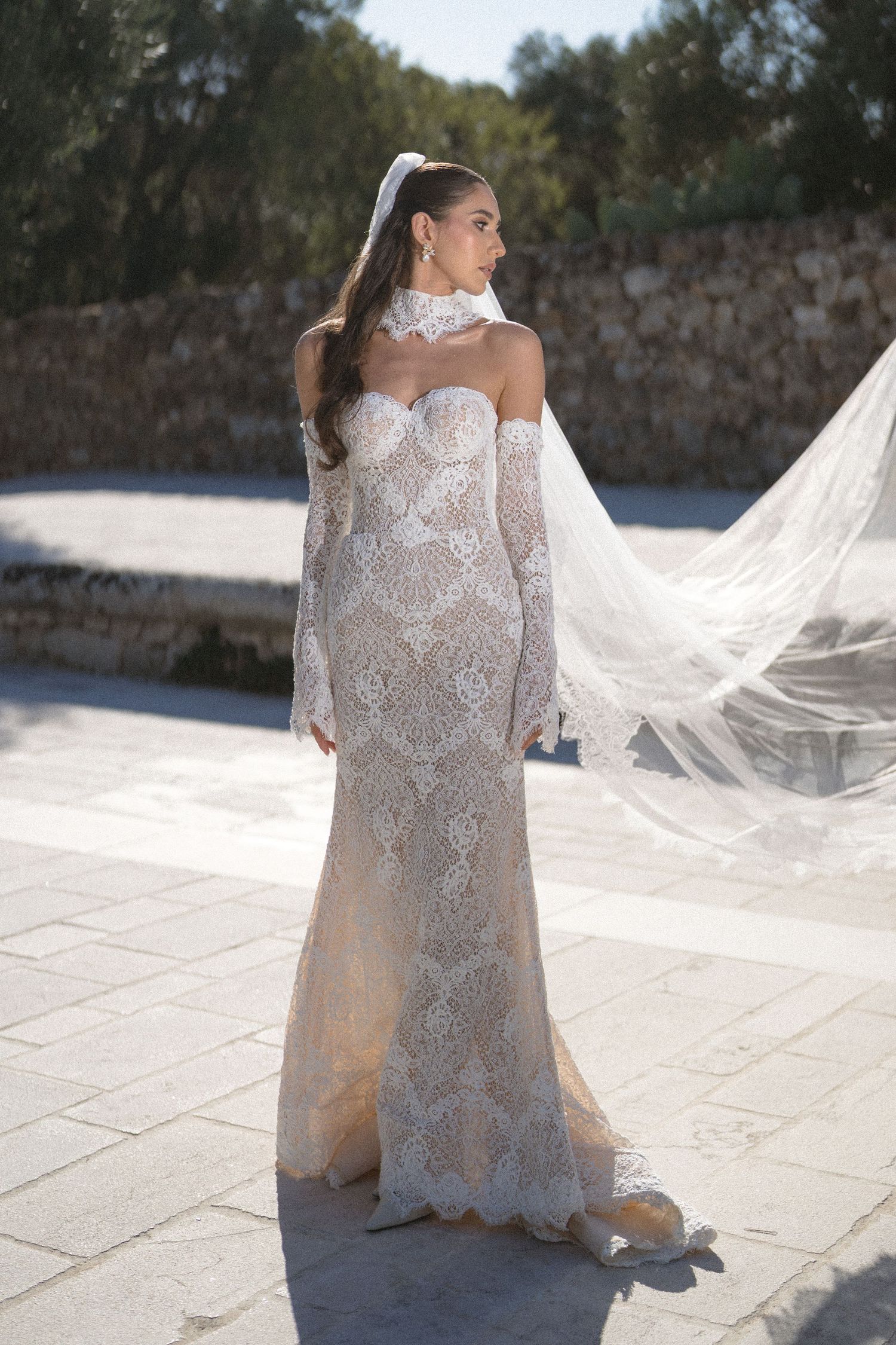 Bride in a fitted lace wedding gown with long sheer sleeves and flowing veil stands on a sunny stone terrace near an old wall.