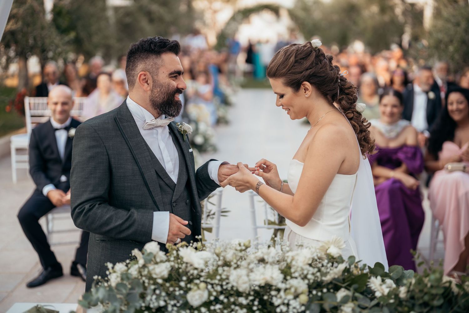 A couple exchanges rings during an outdoor wedding ceremony with white floral decorations and guests looking on.
