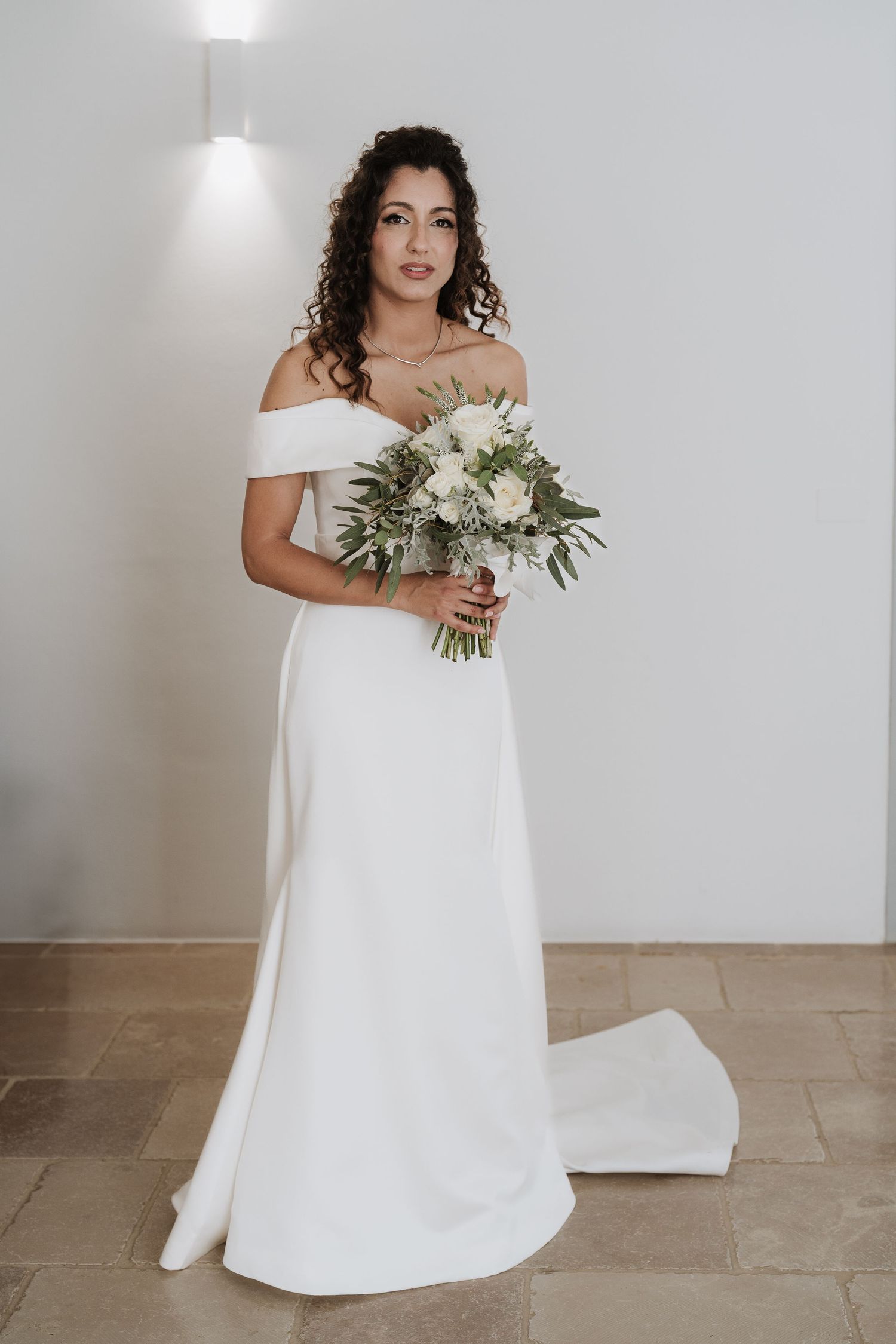 Bride in elegant off-shoulder white wedding gown holds a delicate bouquet of white and green florals.