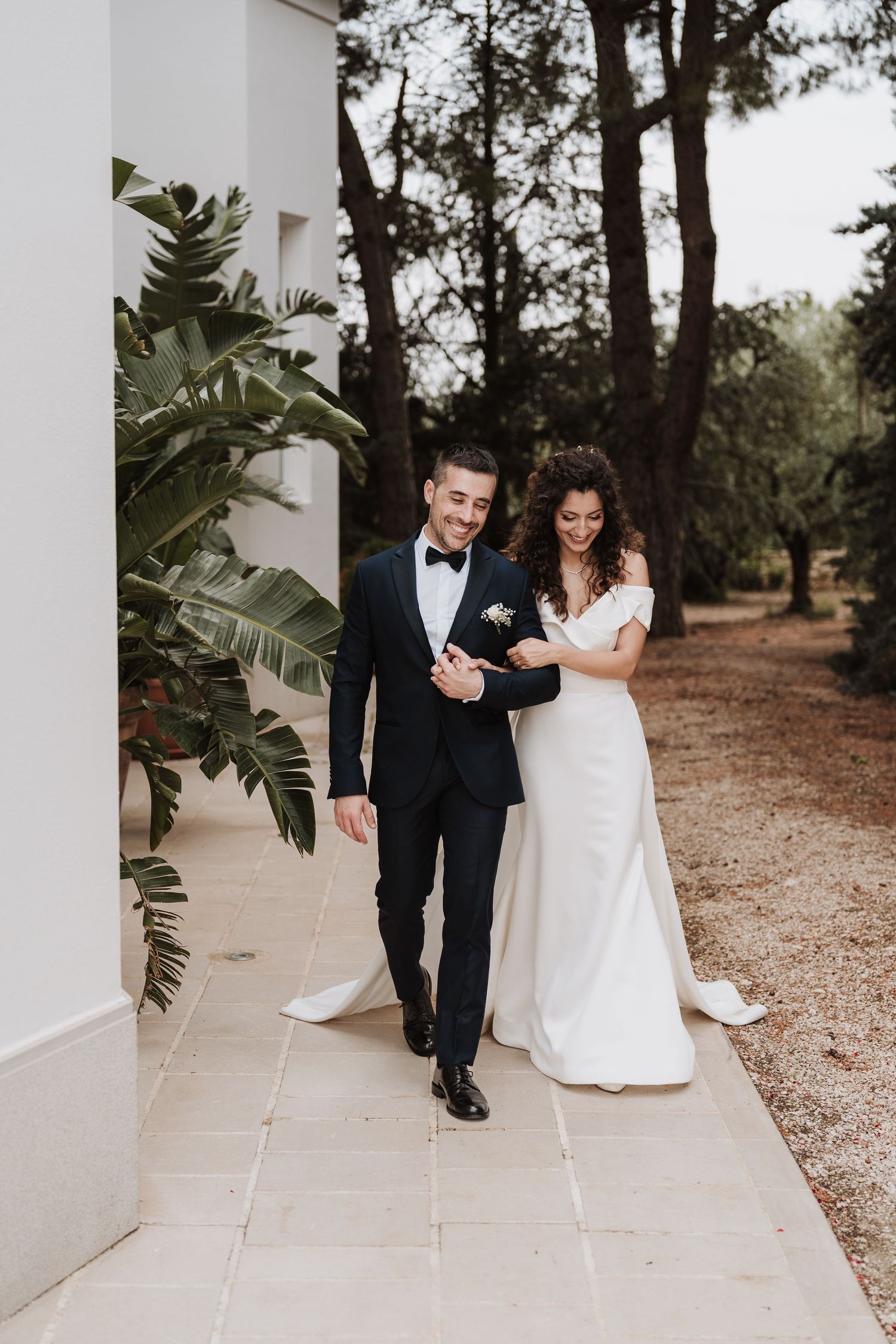 Elegant couple walking along white building pathway with tropical plants and pine trees.