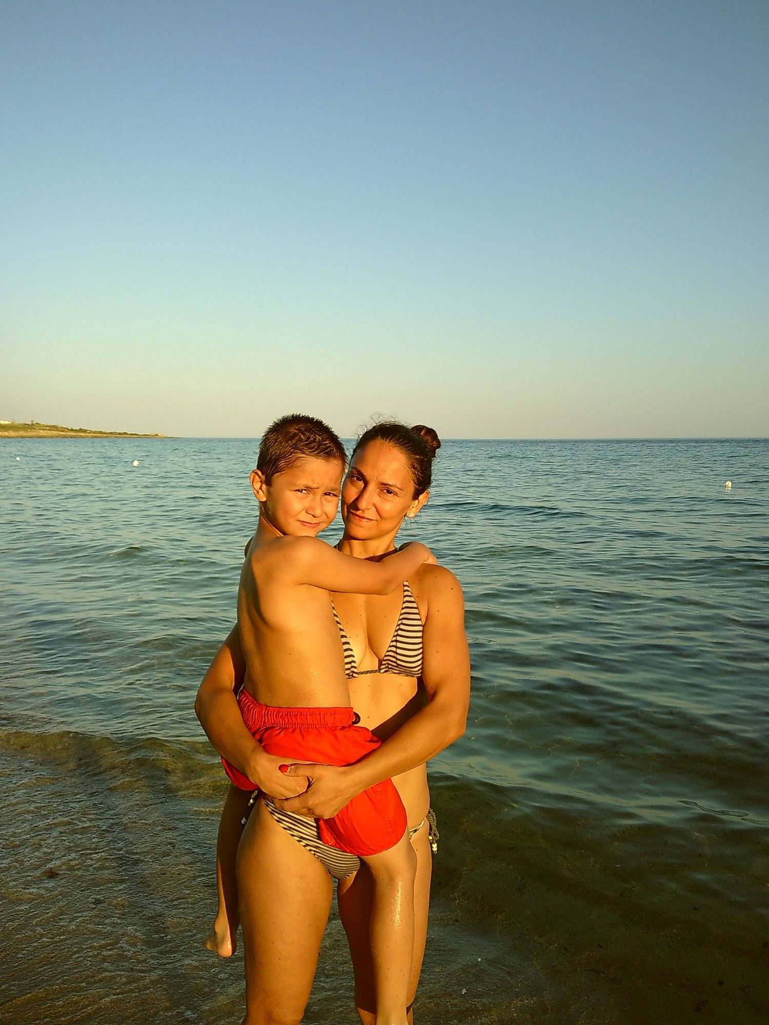 Beachgoers embrace while standing in calm ocean waters on a summer evening.