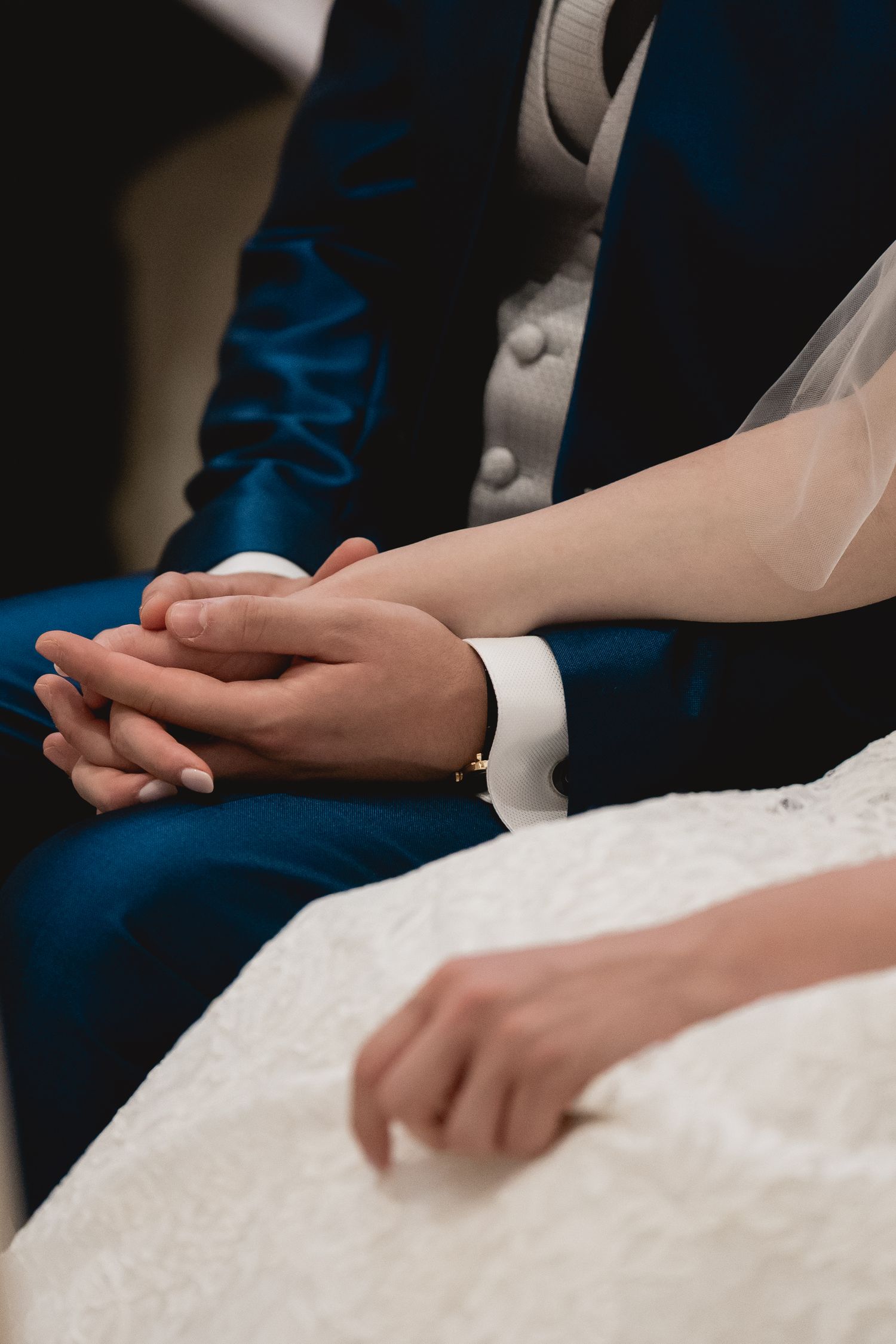 Close-up of clasped hands between a couple in wedding attire showing a navy suit sleeve and white dress.