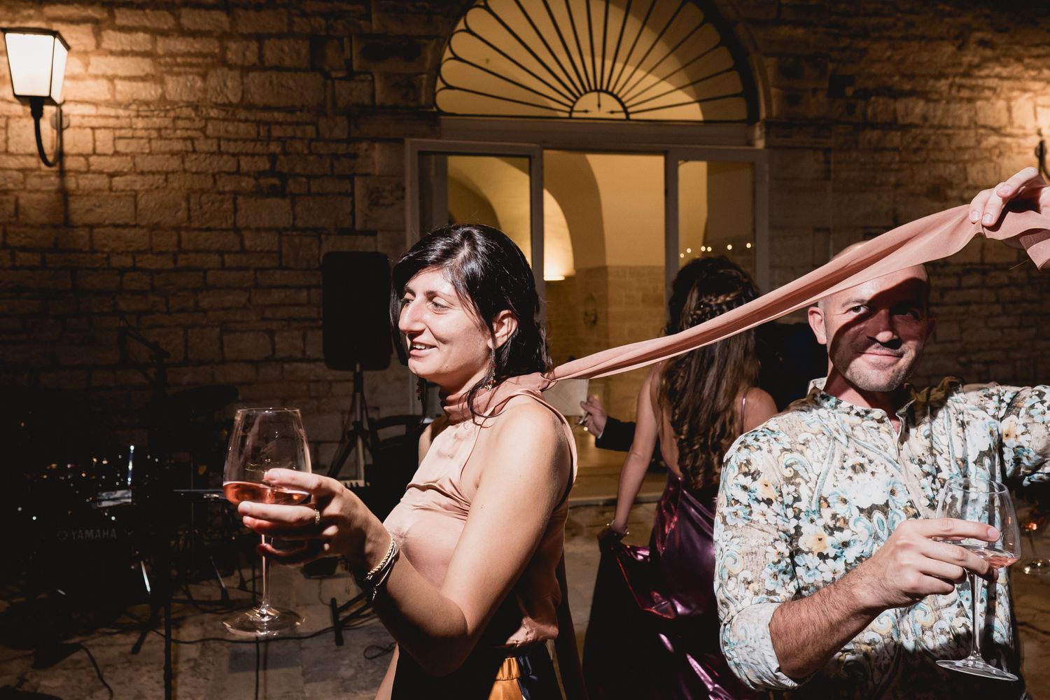 Two people dancing and enjoying drinks at an evening party in front of a stone wall and illuminated doorway.