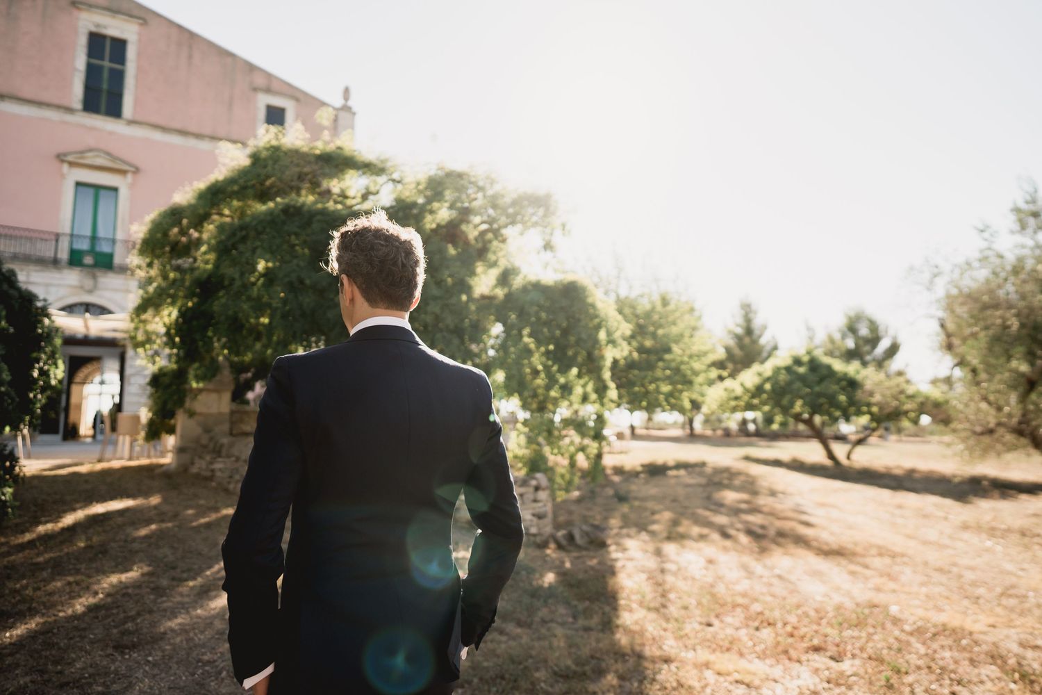 A suited figure stands in contemplation against the backdrop of a pink building and garden landscape.