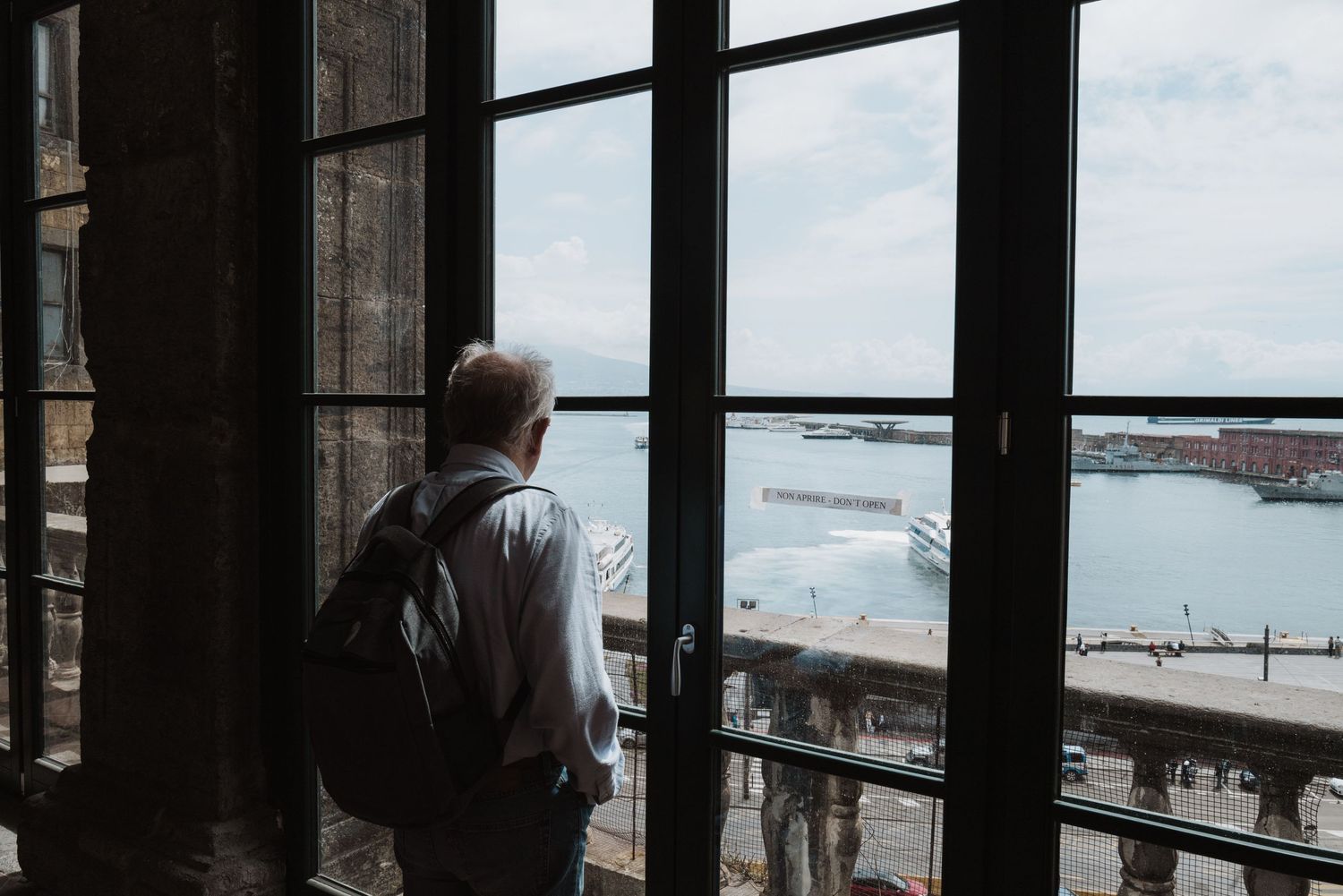 Person standing at window overlooking harbor and city waterfront on cloudy day.
