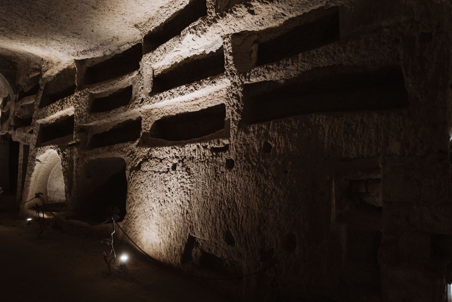 Ancient underground catacombs lit by spotlights revealing carved stone walls and passages.