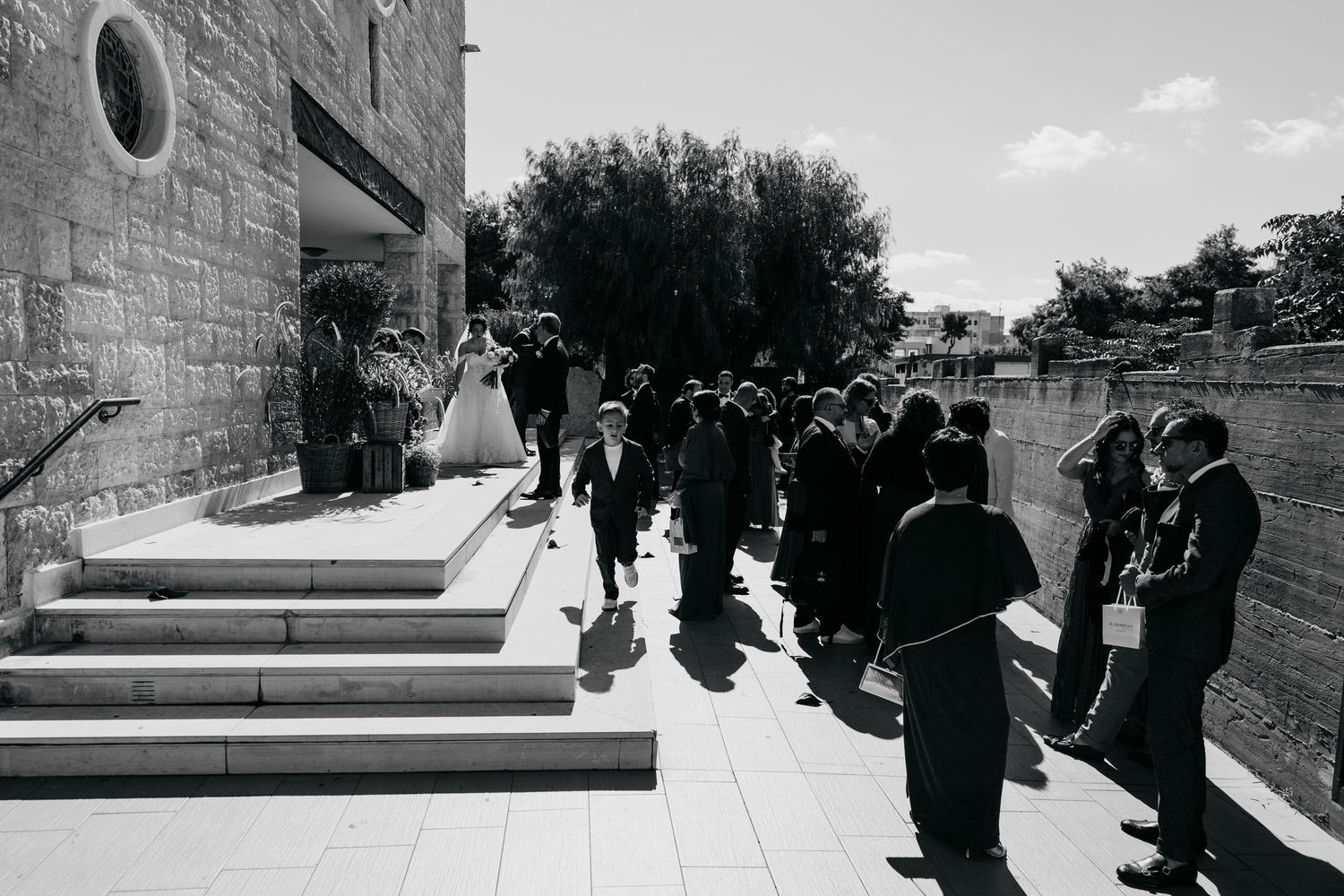 Black and white photo of wedding guests gathered outside a stone church on a sunny day.