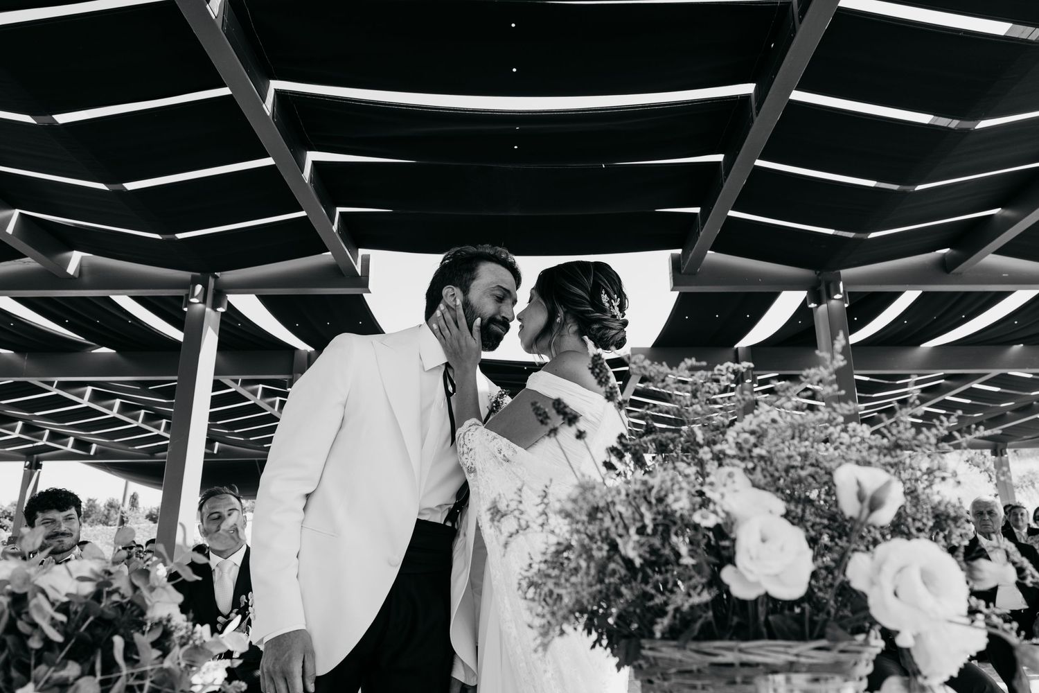 Black and white photo of a couple sharing a kiss at their wedding ceremony surrounded by floral arrangements.