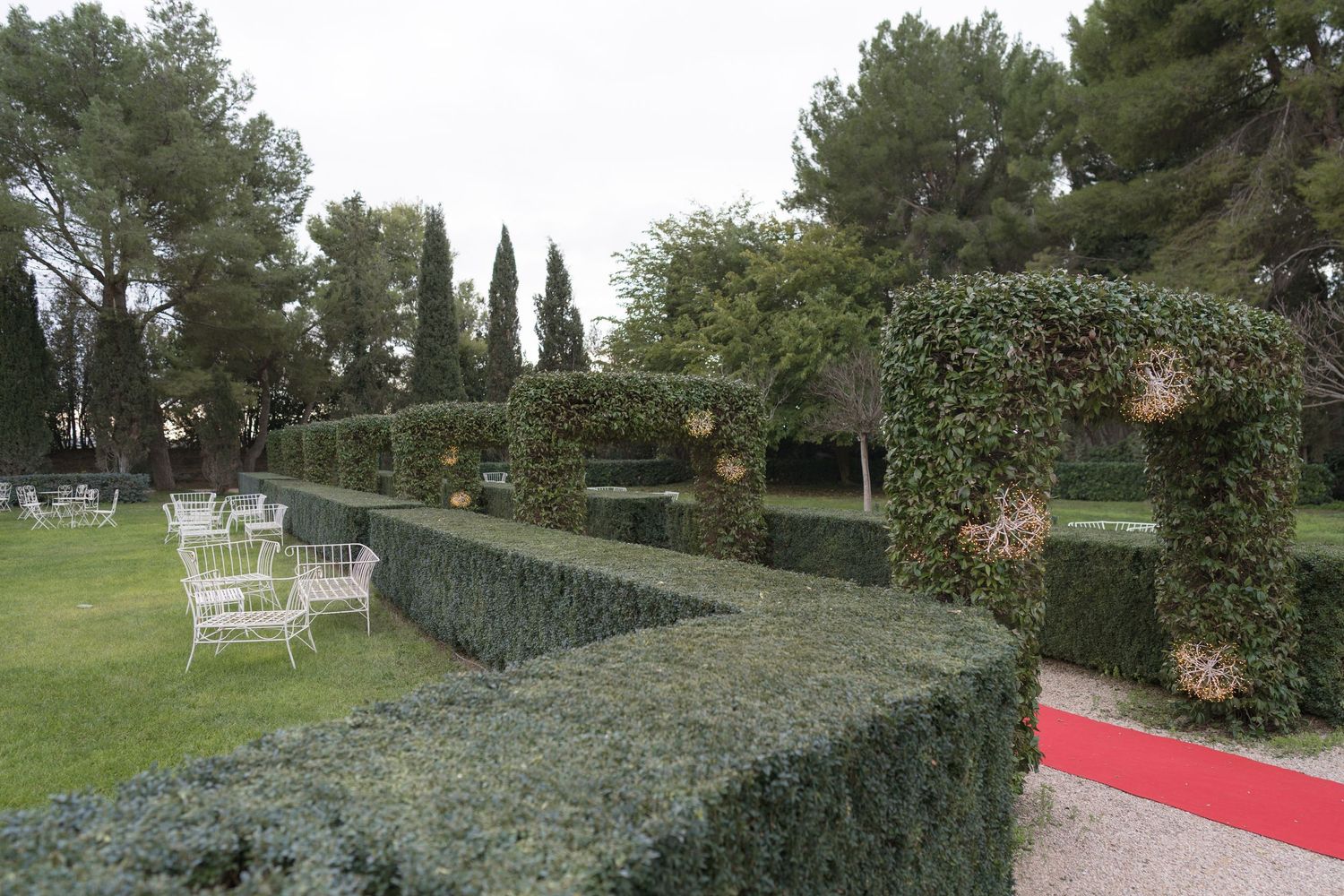 Elegant outdoor wedding venue with manicured hedges, ivy-covered arches, and white chairs on lush green lawn.