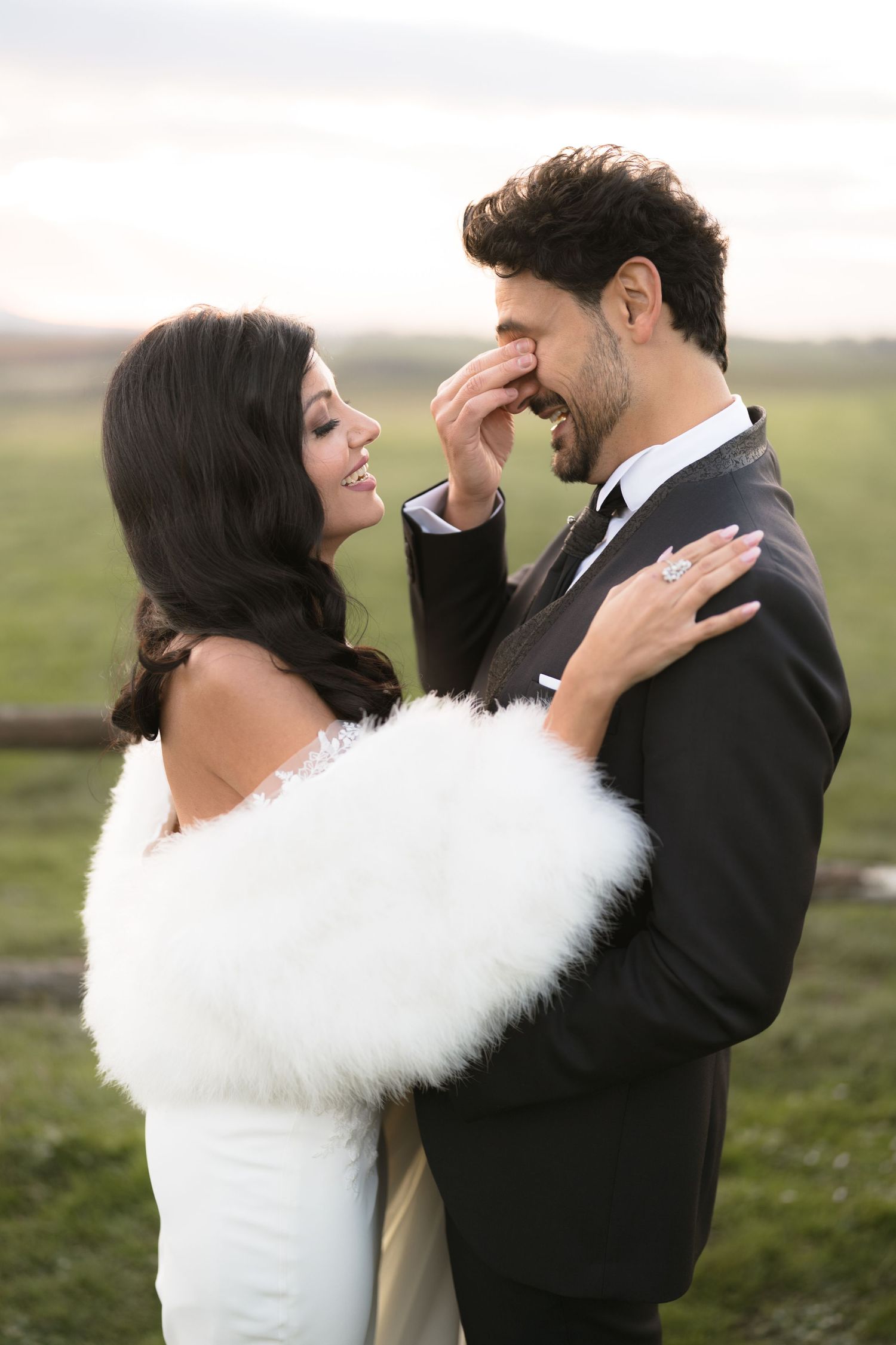 A bride in white dress with fur stole embraces her groom in black tuxedo outdoors in a romantic wedding portrait.