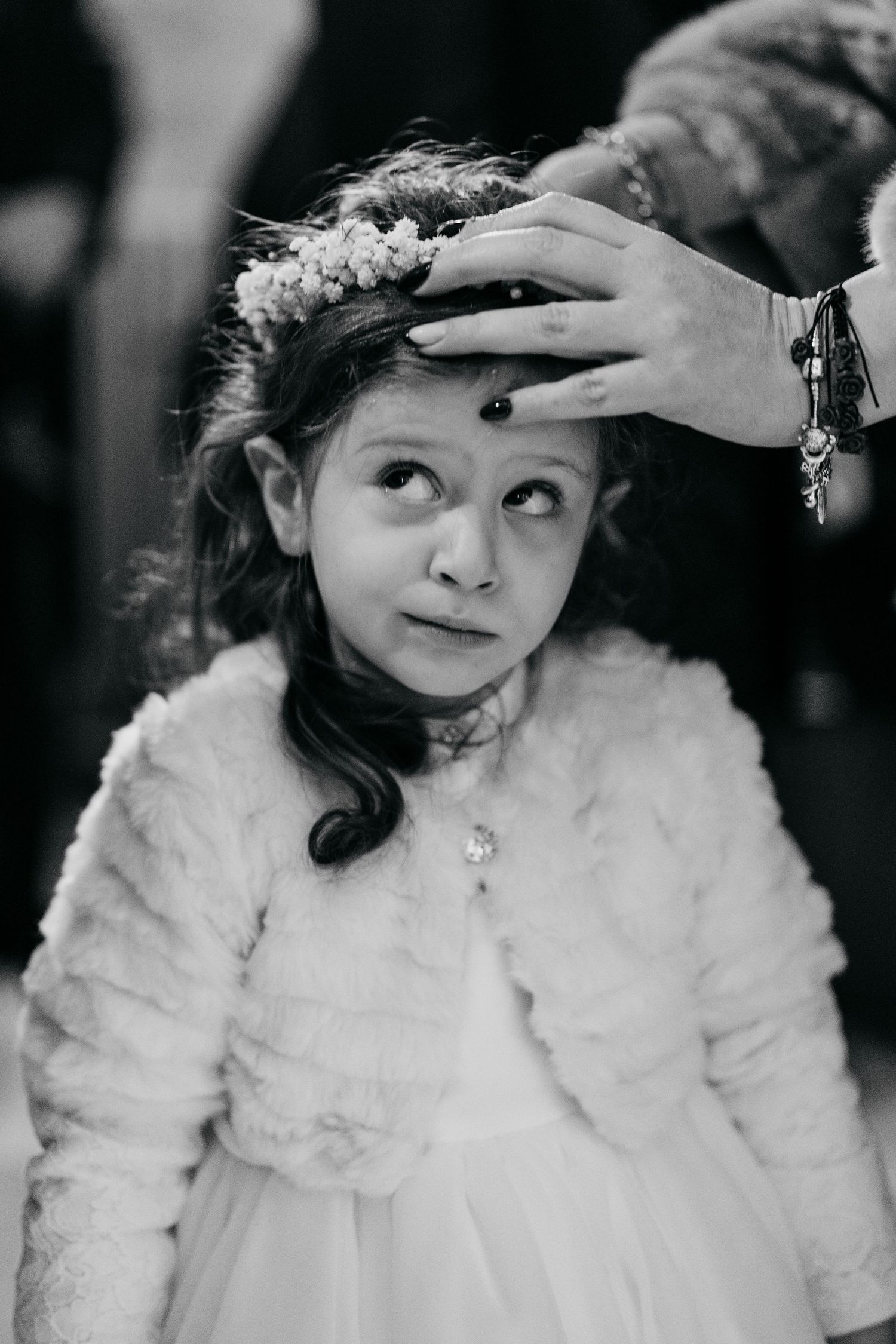 Black and white portrait of young flower girl in white fur jacket with floral crown being adjusted by adult hand.