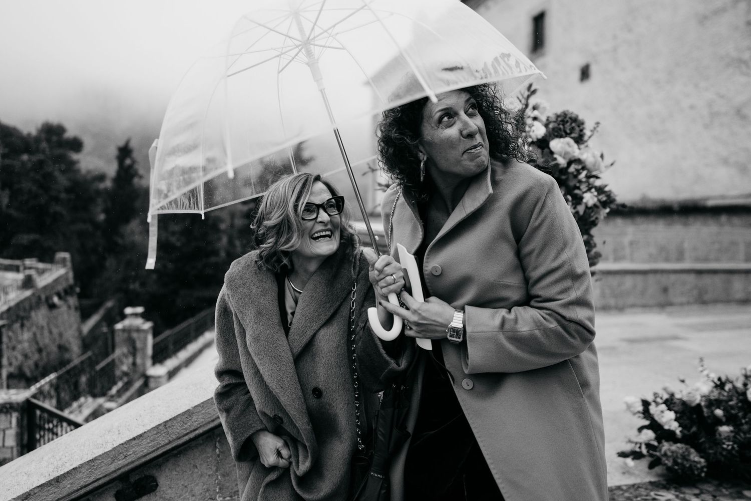 Two women laughing under a clear umbrella on a rainy day outdoors.