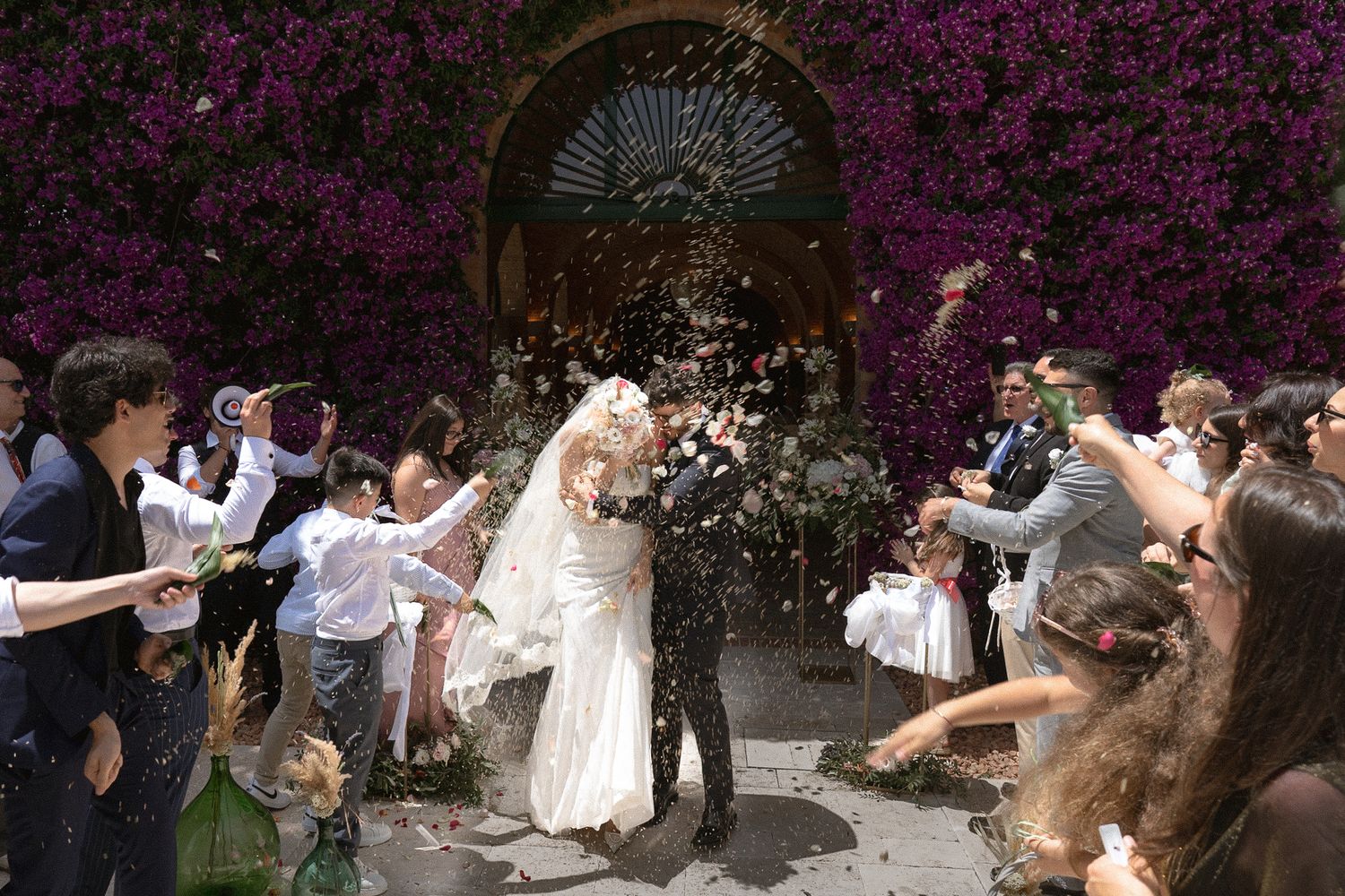 Bride and groom exit wedding ceremony through confetti shower, surrounded by guests, with purple bougainvillea backdrop.