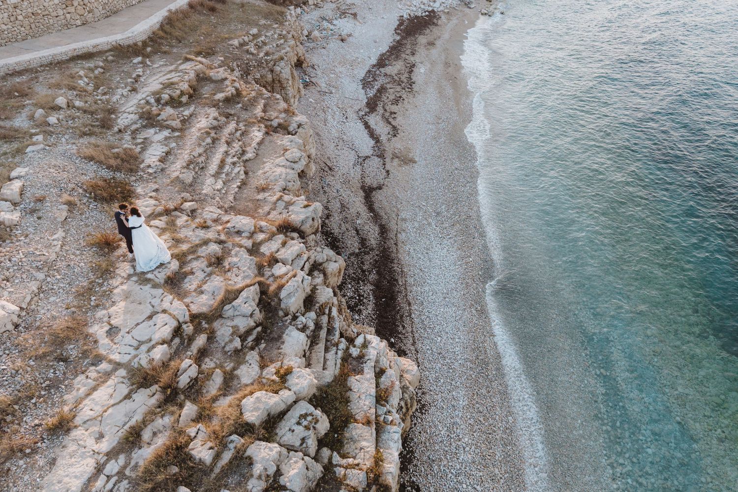 Aerial drone view of a wedding couple standing on rocky coastal cliffs beside turquoise ocean waves.