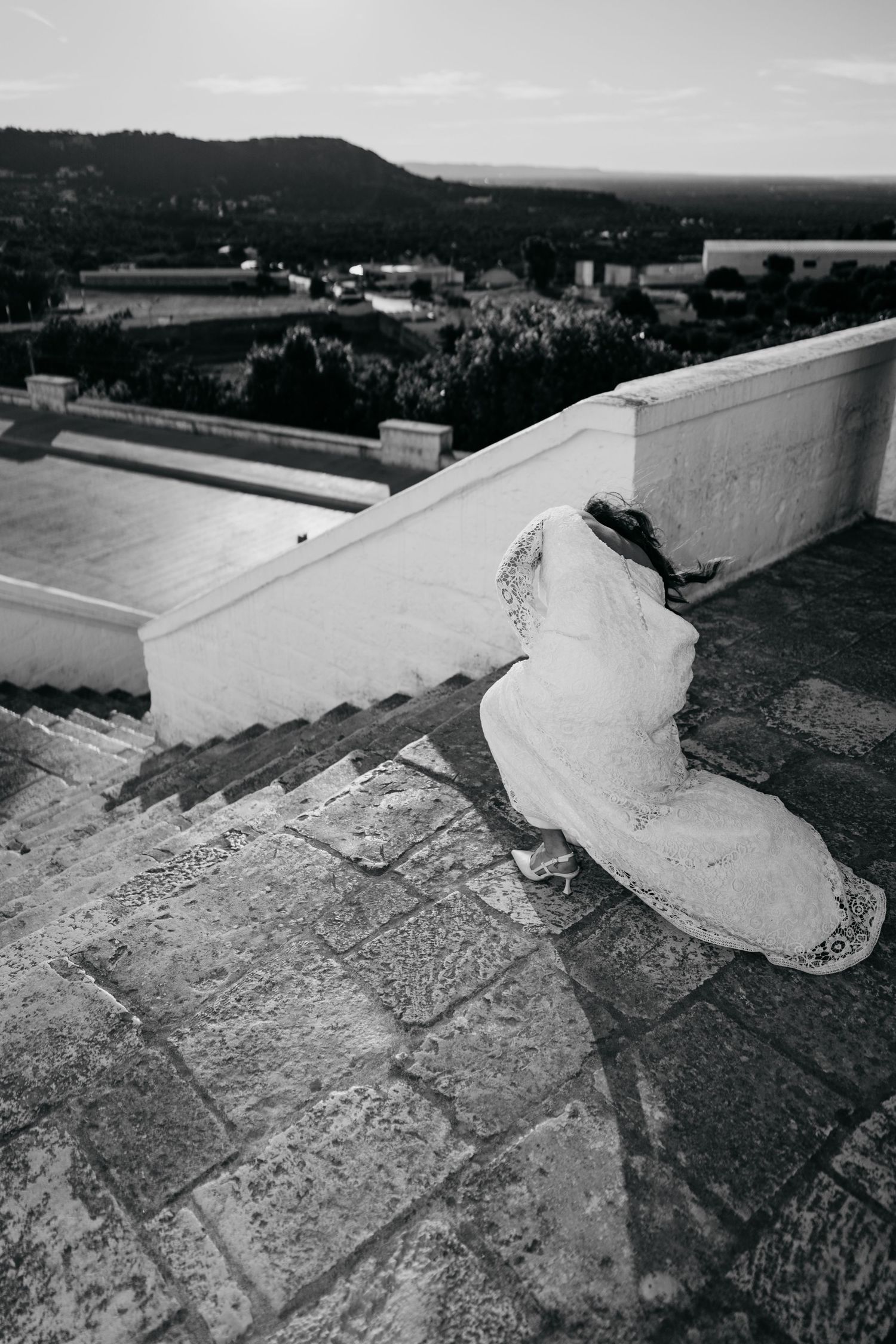 Black and white photo of a bride in a white gown sitting on stone steps with a scenic landscape view.