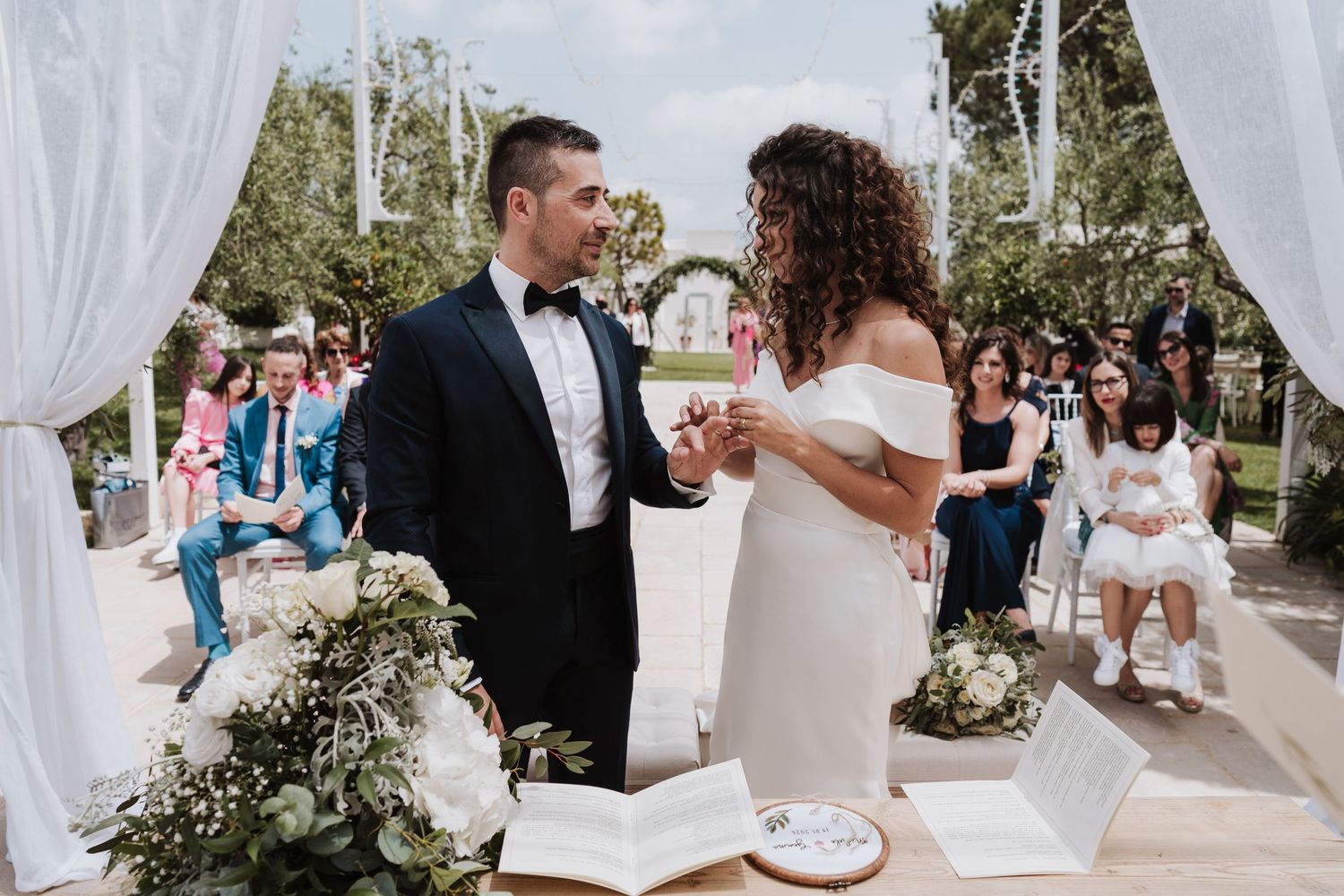 Couple exchanging rings during outdoor wedding ceremony under white draped archway.