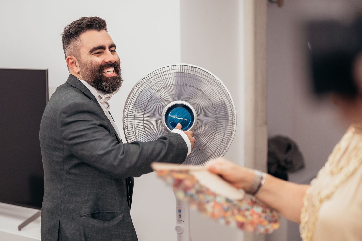 Person in formal suit holding an electric fan while smiling in an indoor setting.