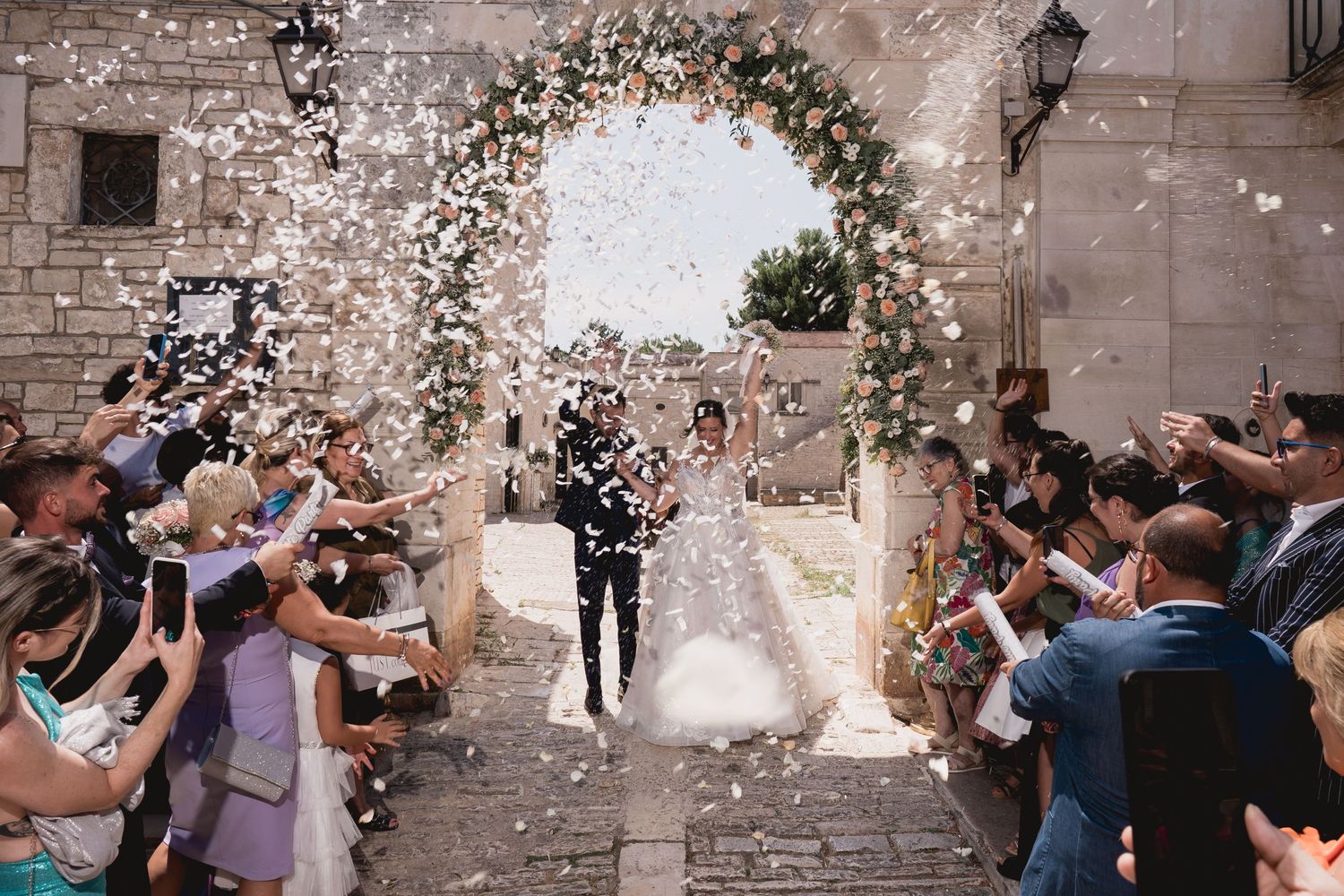 Guests throw confetti at a newlywed couple under a flower-decorated stone archway.