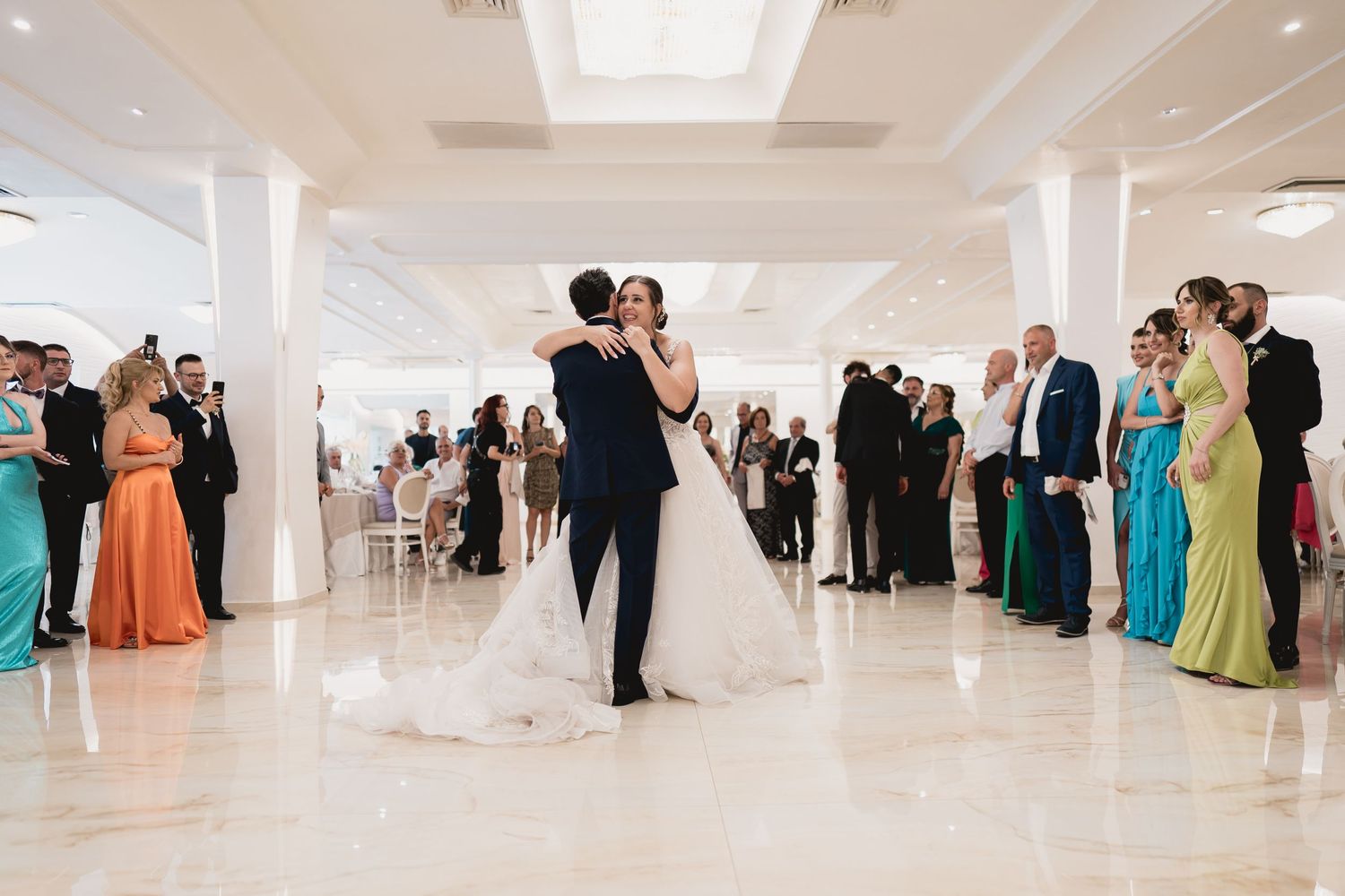 A newlywed couple dances while guests in colorful formal attire watch during their first dance.