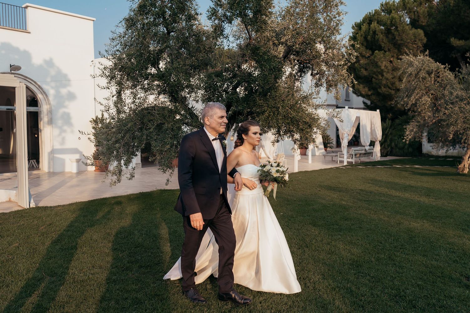 Couple walking together on grass with white building and trees in background.