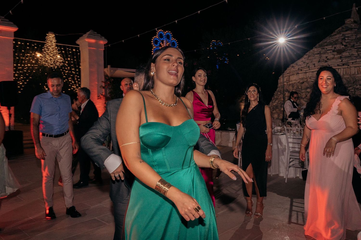Wedding guests dance and celebrate on an outdoor patio illuminated by string lights and ambient red lighting.