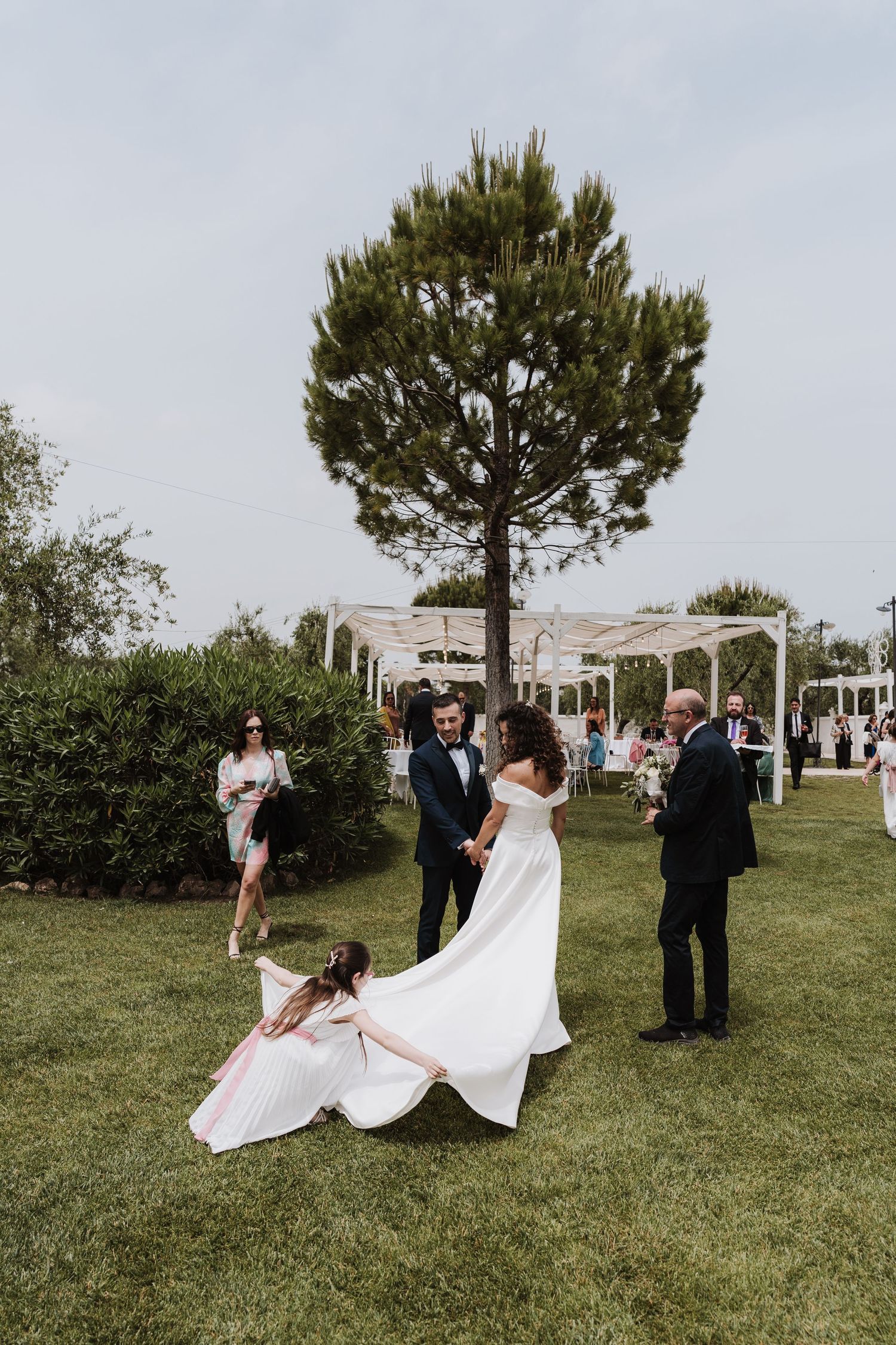 Outdoor wedding scene with flowing white dress and guests gathering under a pine tree.