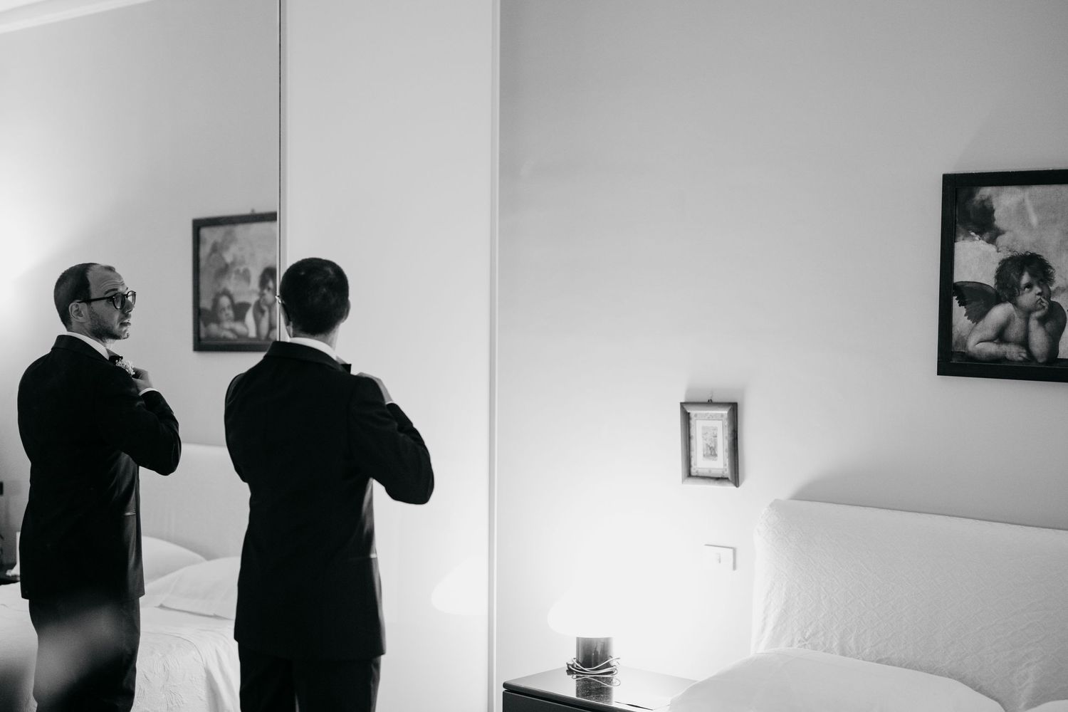 Two people in suits prepare for a formal event while looking in a mirror in a white bedroom.