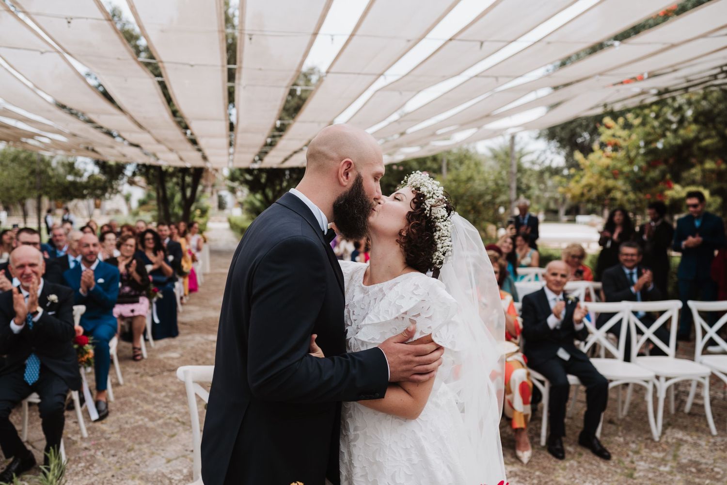 Romantic wedding moment under white canopy as guests look on from white garden chairs.