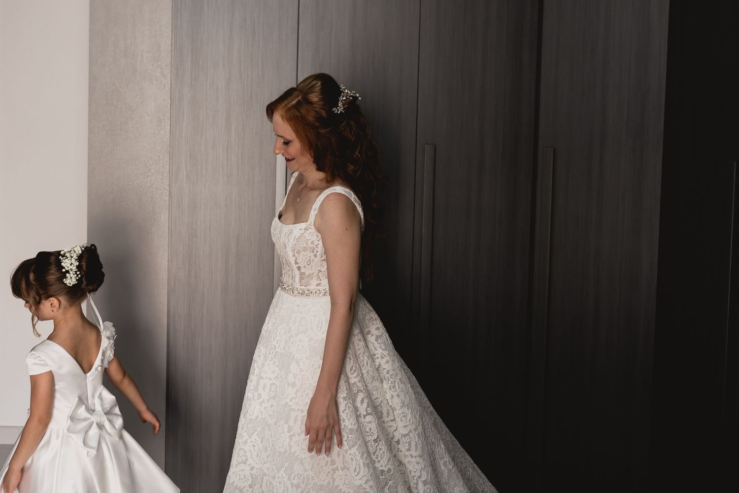 Two people in elegant white dresses stand against a dark wall in a dreamy wedding photo.