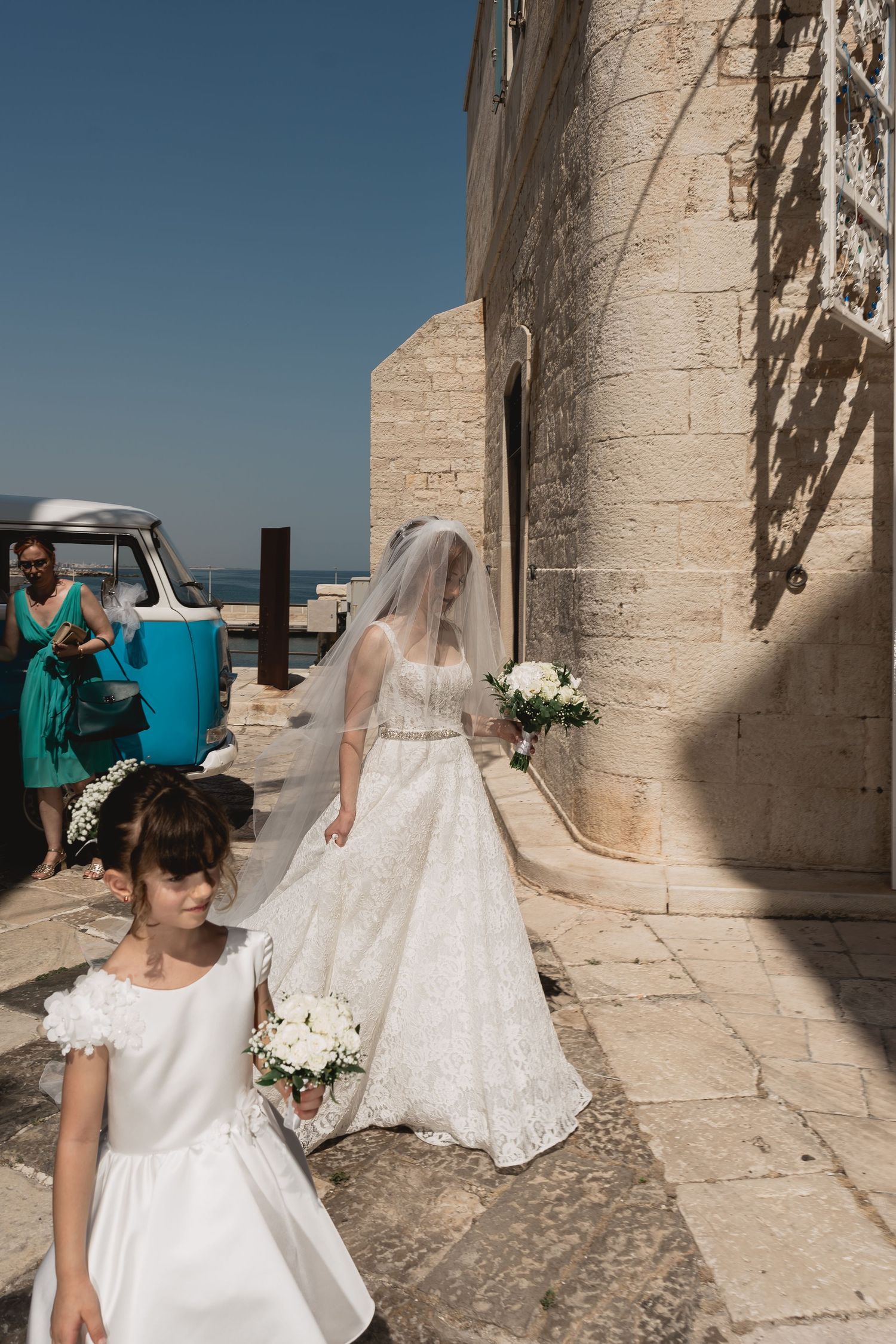 Wedding party walks along stone pathway with classic car and Mediterranean architecture in background.