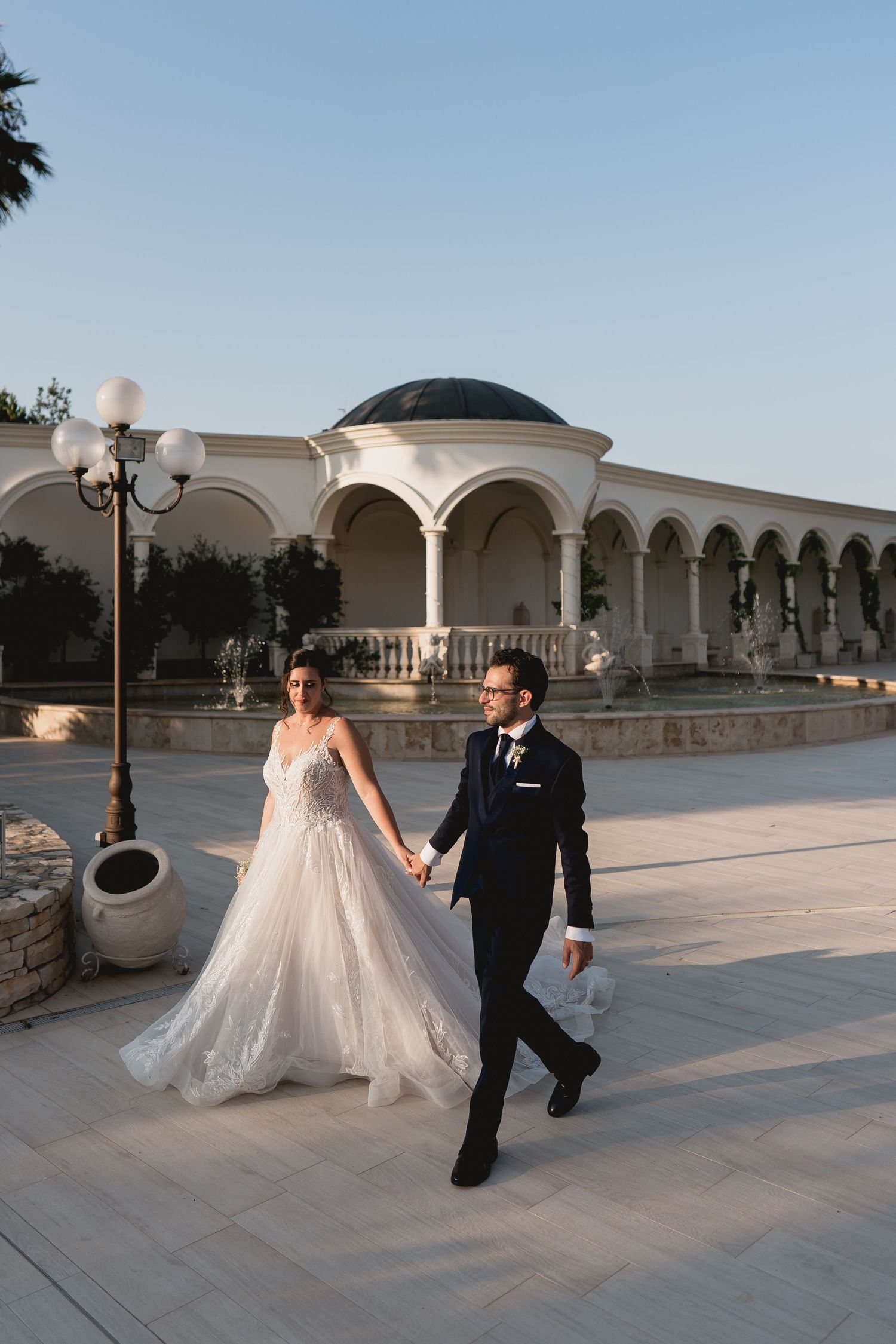A couple in wedding attire walk hand-in-hand across a courtyard with elegant white arched columns and decorative lamp posts.