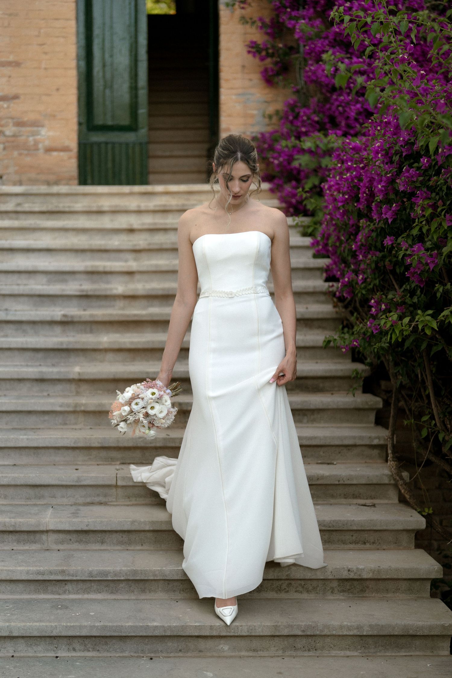 Bride in white dress descending stone steps with purple flowers in background.