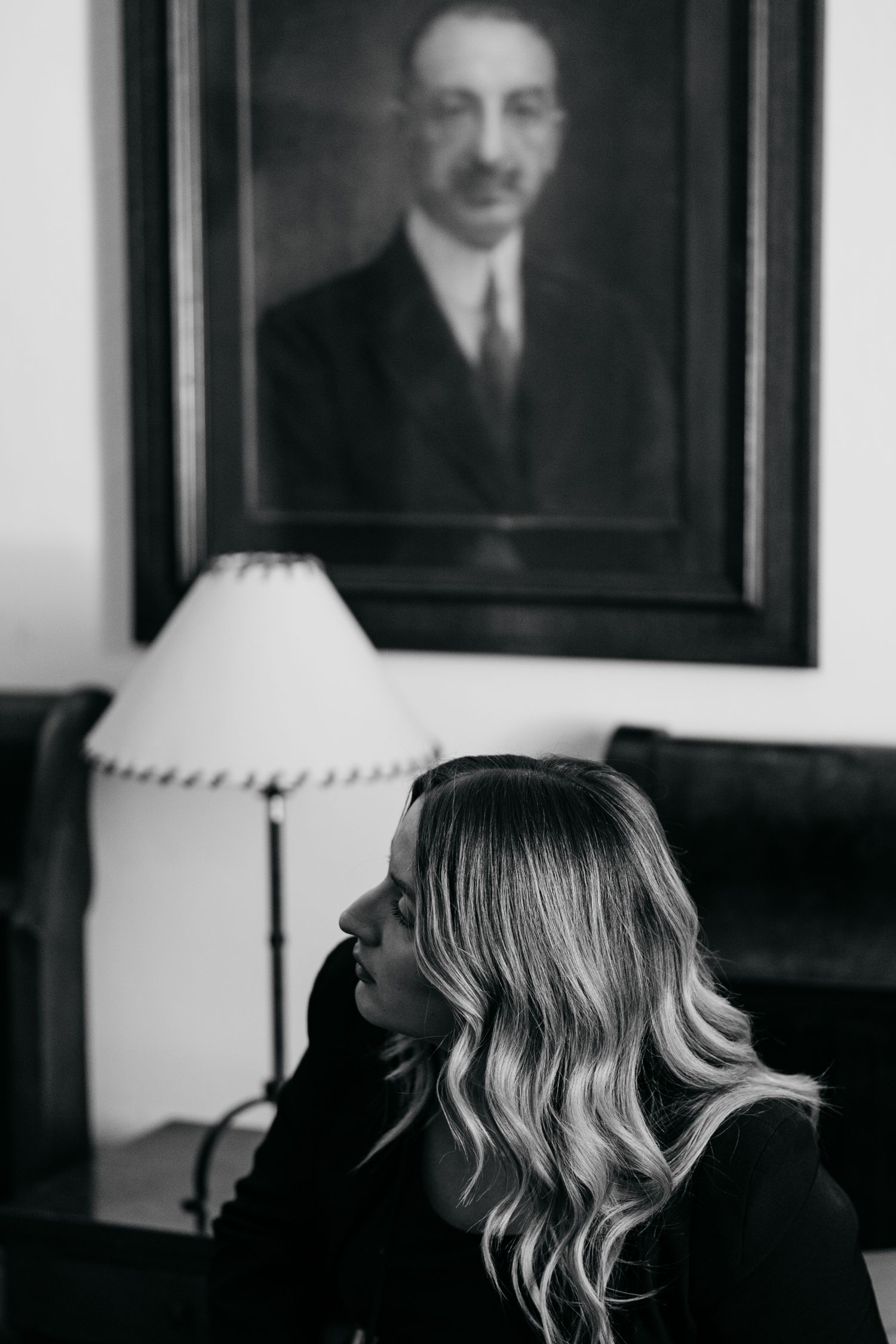 A black and white photograph shows a series of framed portraits hanging on a wall with a lamp in the corner.