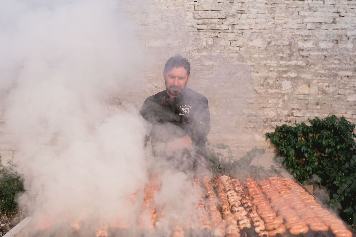 Chef grilling and smoking meat on large outdoor barbecue against brick wall.