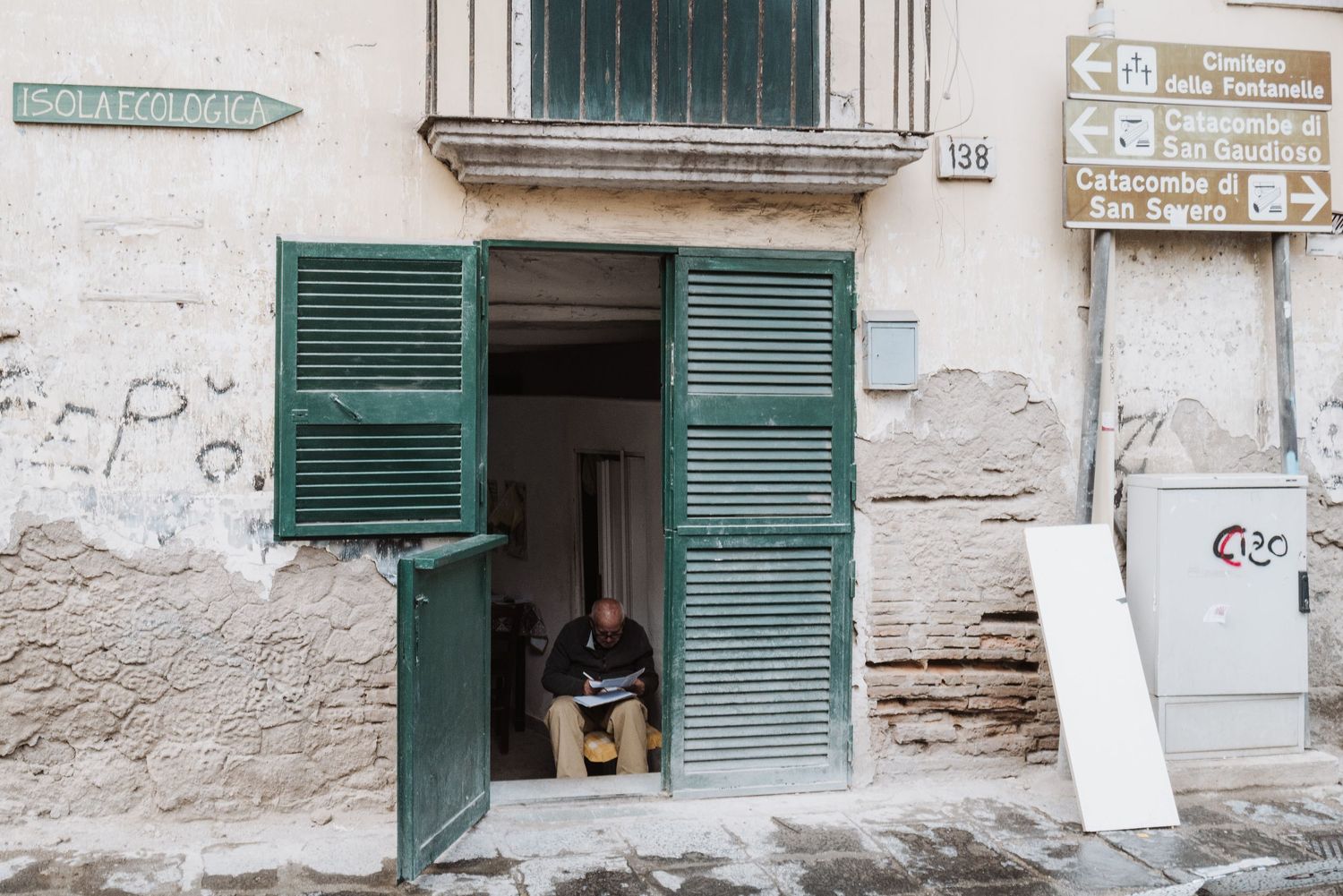 Dog sitting in a doorway with green shutters on a weathered building facade.