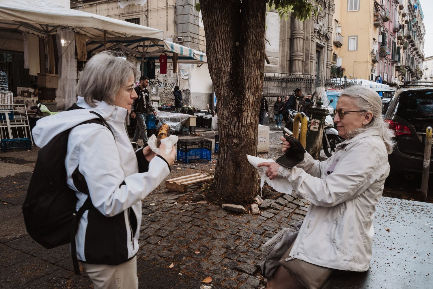 Two tourists take photos on a cobblestone street under a large tree in an urban setting.