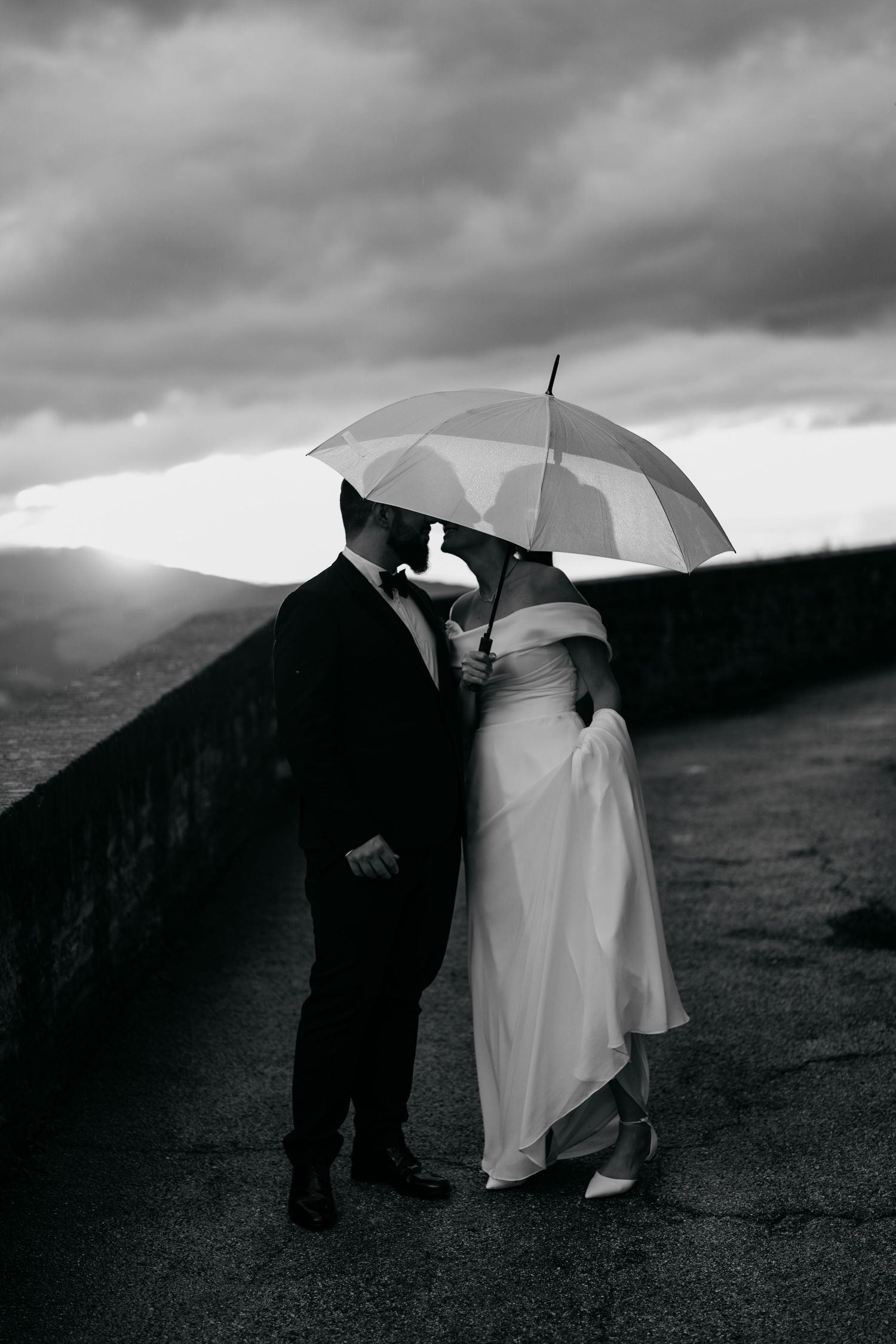 Dramatic black and white photo of wedding couple with umbrellas against moody mountain backdrop.