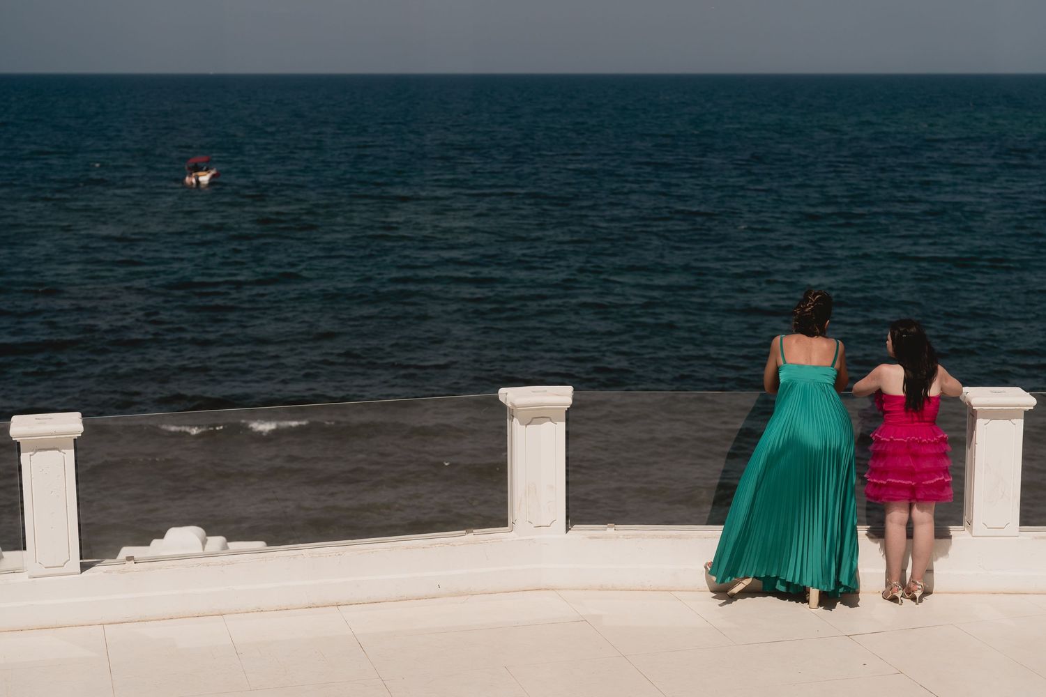 Two figures stand on a white balcony overlooking a dark ocean at dusk.