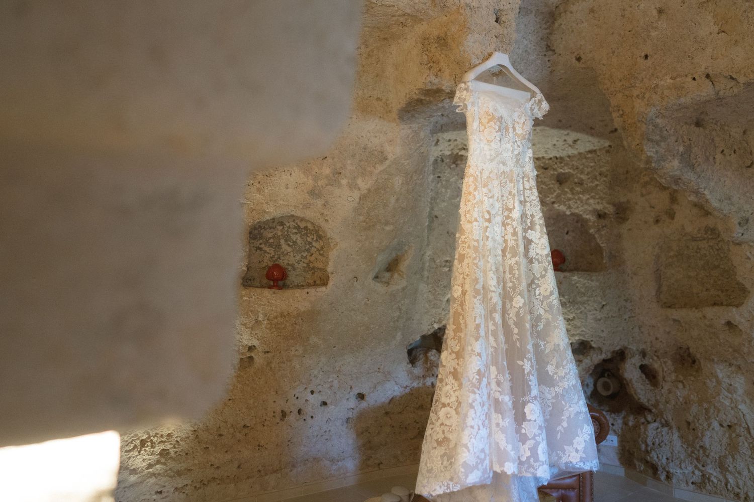 White lace wedding dress hangs elegantly in a rustic stone archway with natural lighting.