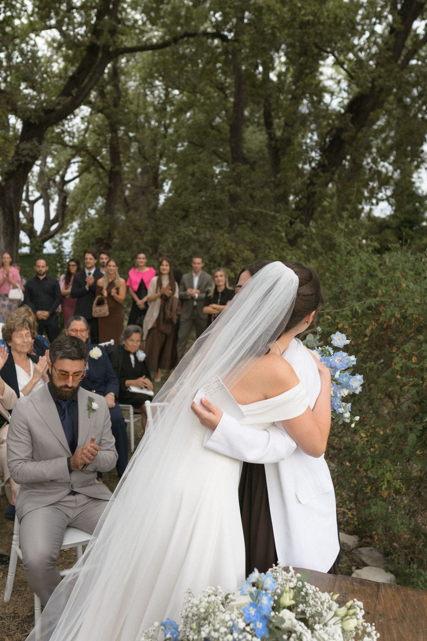 A wedding ceremony takes place outdoors with guests watching as a couple shares a romantic moment under a white umbrella.