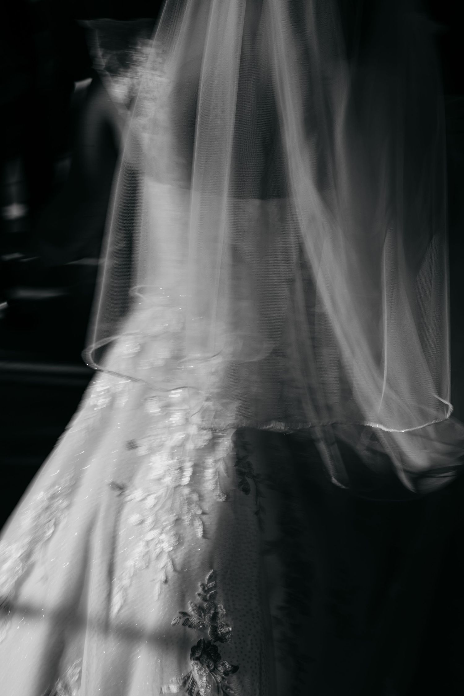 Artistic black and white close-up of bride's lace wedding gown and flowing veil in soft light.