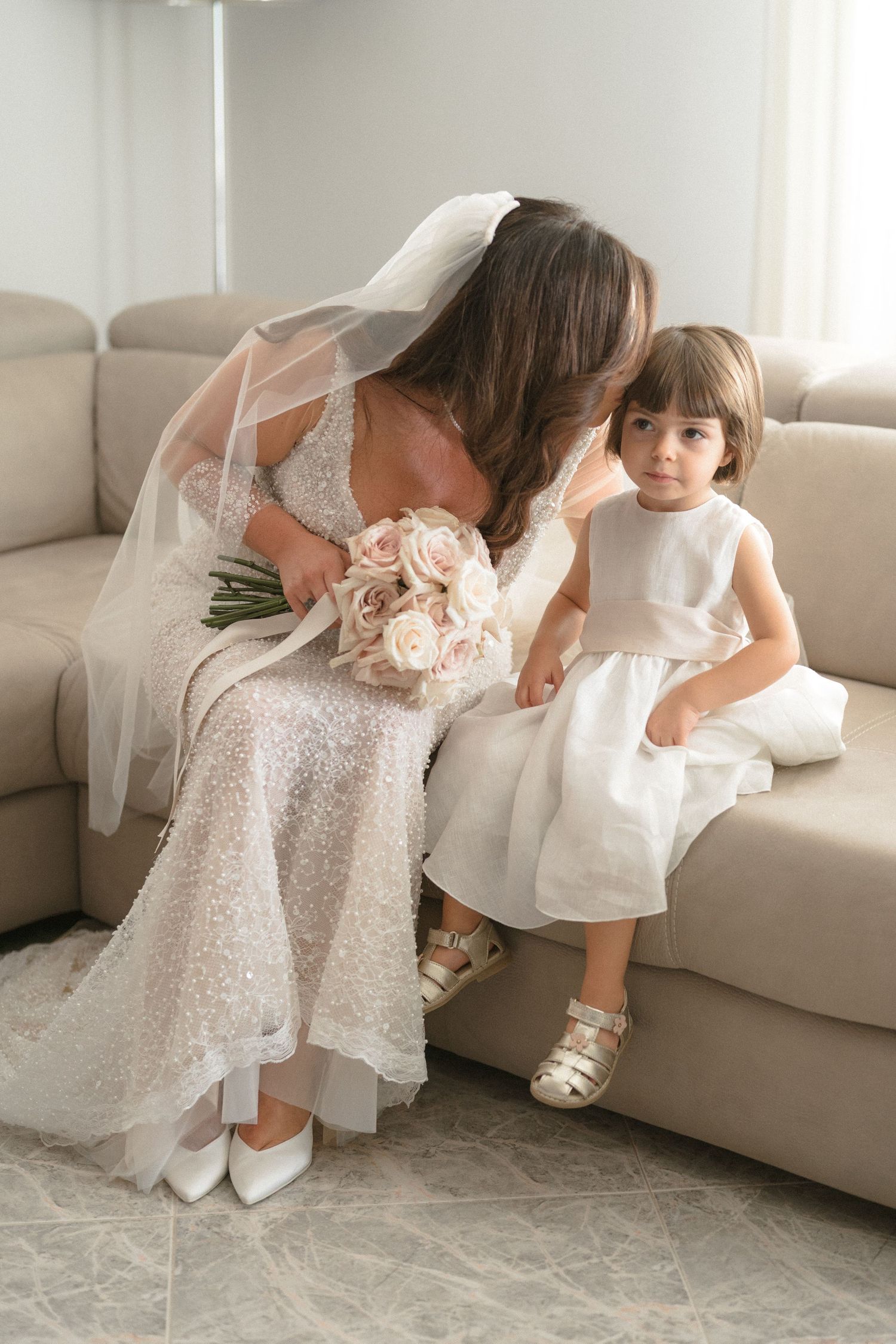 Two children in white dresses sit together on a light colored couch in an intimate indoor setting.